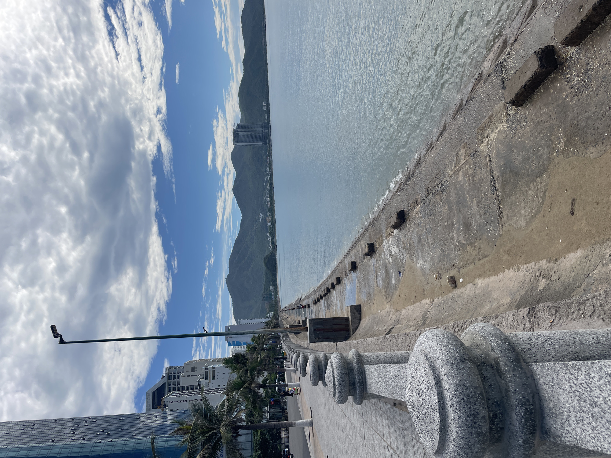 The image captures a vibrant coastal scene in Bac Nha Trang Ward, Vietnam, featuring a promenade, the ocean, and distant mountains under a partly cloudy sky. The perspective is from the promenade, looking along the coastline.
In the foreground, a paved pedestrian walkway stretches into the distance, bordered on the right by a decorative, speckled grey stone balustrade with prominent cylindrical elements. Several slender palm trees line the promenade, and a tall streetlight with a modern LED fixture stands on the left. A few indistinct figures are visible on the walkway, suggesting people are strolling or commuting.
To the right of the promenade, a concrete sea wall or embankment separates the walkway from the calm, light blue-grey ocean. The wall appears somewhat weathered, with large rectangular blocks extending into the water near the shore. The ocean surface shows subtle ripples and small waves breaking gently against the base of the wall.
In the midground and background, modern multi-story buildings, likely hotels or residential complexes, are scattered along the coastline, nestled against the foothills of large, dark green mountains. One building clearly displays "BOSTON" vertically, and another partially shows "KING" or similar text. The mountains form a dramatic backdrop, rising steeply from the coast.
The sky is bright and partly cloudy, with a mix of fluffy white cumulus clouds and some larger, darker grey formations. The time of day appears to be daytime, likely late morning or early afternoon, given the clear visibility and brightness. The overall atmosphere conveyed is peaceful and inviting, typical of a coastal resort or city area.