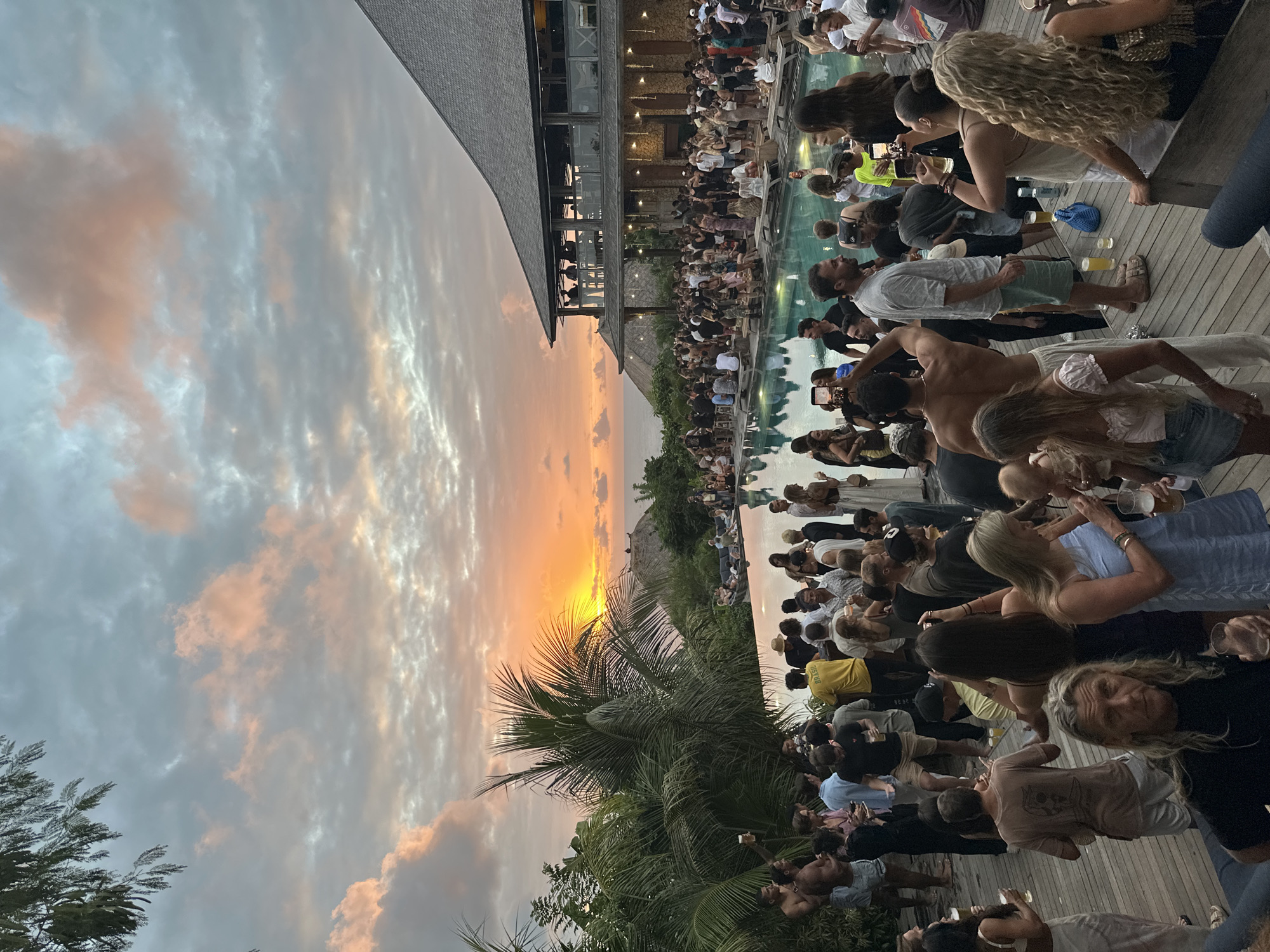 This image depicts an outdoor gathering at a resort or villa in Kuta Selatan, Indonesia, during sunset. The scene is set on a wooden deck surrounding an infinity pool, with a large, multi-story building in the background that appears to be part of the resort's architecture. Lush tropical vegetation, including palm trees, is visible around the pool and property, suggesting a coastal or island location.
The sky is filled with dramatic sunset clouds, painted in hues of orange, pink, and gray, with the sun low on the horizon, casting a warm glow over the scene. The ocean is visible in the distance.
A large group of people, estimated to be over fifty individuals, are scattered across the deck, engaged in various activities. Many are holding drinks, conversing, and socializing. Some individuals are standing, while others are seated on wooden benches and low tables. The attire is casual, typical for a beach or resort setting, with many people in swimwear or light summer clothing.
Several individuals are seen holding smartphones, suggesting they are capturing the sunset or the event. The atmosphere appears lively and festive, with a sense of relaxed enjoyment. The pool itself is illuminated, reflecting the ambient light. The resort building features a distinctive thatched roof and open-air balconies, characteristic of tropical architecture.