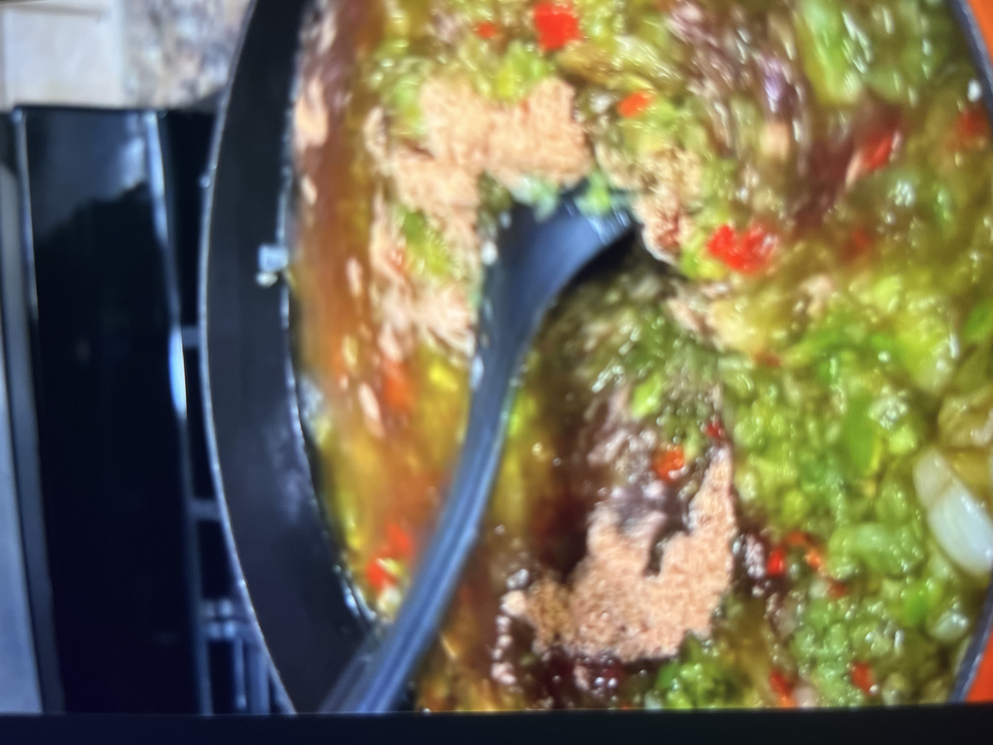 The image shows a close-up, blurry view of a black cooking pot filled with a liquid mixture containing chopped green vegetables, red pepper pieces, and some reddish-brown granular material. A black utensil is partially submerged in the mixture, appearing to stir or serve it. The background is out of focus and shows what looks like a stovetop and countertop, suggesting a kitchen environment. There is no indication of the city of Abuja, Nigeria, in the image.