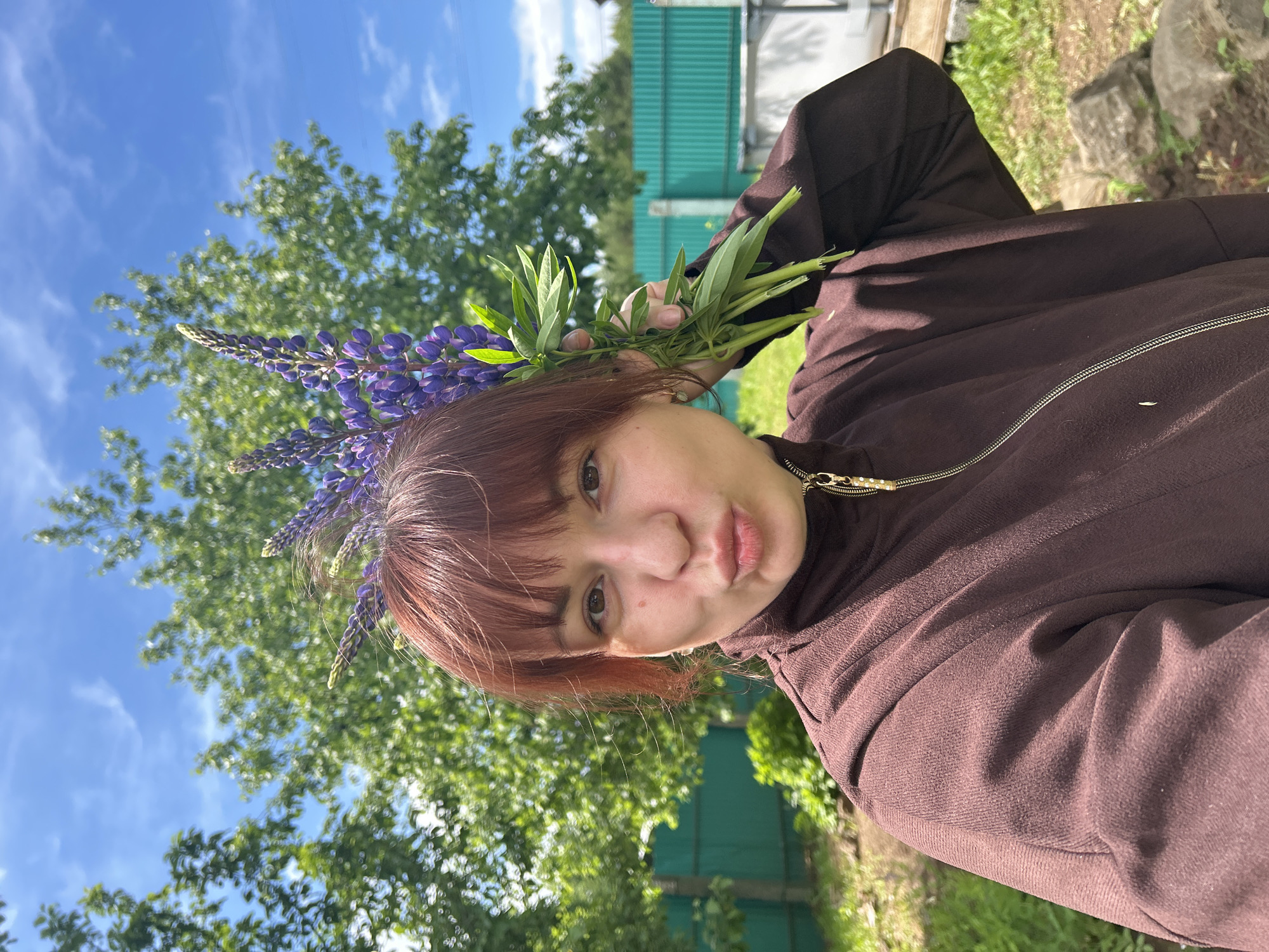 A young woman with reddish-brown hair holds a bouquet of purple lupine flowers to her head, against a backdrop of green foliage and a blue sky with wispy clouds. The woman has fair skin and is wearing a brown jacket with a gold zipper. The setting appears to be outdoors, with a green fence and trees visible in the background. The location is implied to be Фоки, Russia.