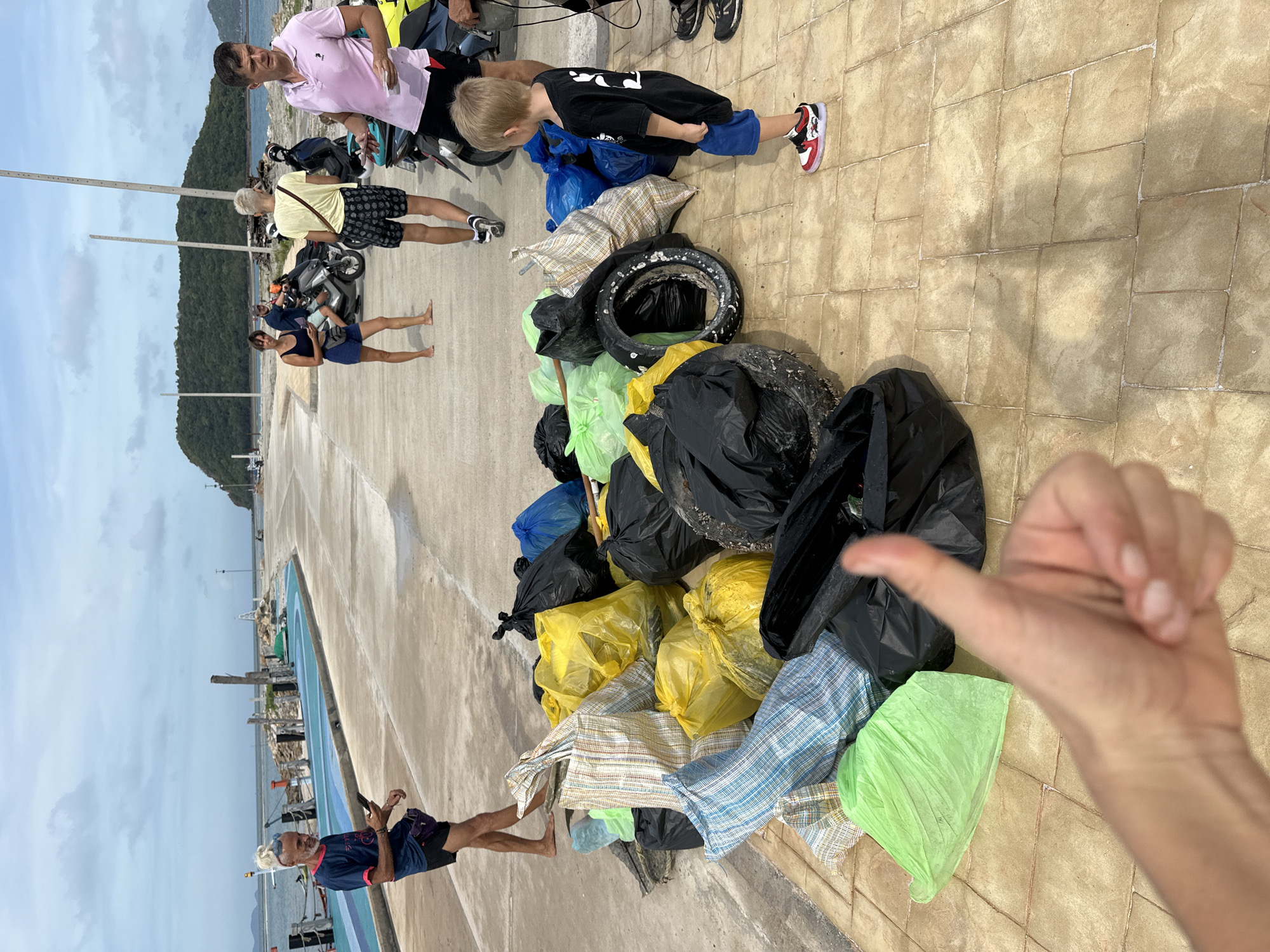 On a paved walkway in Ko Pha Ngan, Thailand, a pile of trash bags, including black, yellow, green, and blue plastic bags, along with woven sacks and two car tires, is stacked on the ground. Several people are visible in the background. A man in a dark blue t-shirt and black shorts stands to the left, looking at his phone. Behind him, a woman in a black tank top and blue shorts walks away. Further back, a man in a yellow t-shirt and patterned shorts is seen with a group of people near motorcycles. A young boy in a black t-shirt and blue shorts stands near the trash pile, facing away from the camera. A hand with a thumb up is in the foreground, partially obscuring the trash. The background features a body of water, a pier with boats, and a lush green hill. The sky is cloudy.