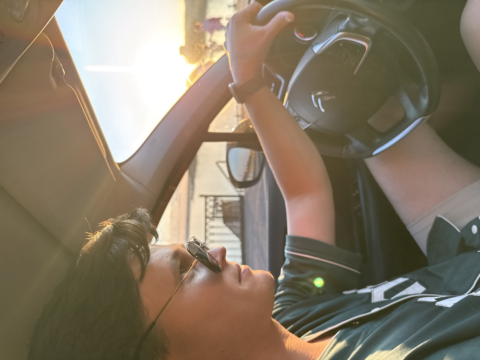 An internal view of a modern Citroën car shows a young adult male, facing right, in the driver's seat at sunset. He has dark, wavy hair and fair skin, wearing dark-tinted aviator-style sunglasses over what appear to be clear-lensed glasses with thin metal frames. He is dressed in a dark green athletic jersey with white trim and partially visible white numbering/lettering on the front. A dark-colored smartwatch is on his left wrist, which rests on the steering wheel, identifiable by the Citroën double-chevron logo and silver accents. Warm, golden sunlight, originating from the right side of the frame, illuminates the car's interior and the driver's profile, casting a soft glow. Through the windshield, a wide body of water is visible in the background, indicative of the Izhevsk Pond. A dark metal fence or railing runs along the water's edge, suggesting a promenade or embankment. Beyond the fence, blurry figures of people are discernible, with one figure prominently in a purple top, signifying a public recreational area. The sky above the horizon is a bright golden-orange, transitioning to light blue overhead, confirming the golden hour. A prominent vertical light flare is present on the upper right side of the windshield. The scene takes place at the Izhevsk Pond in Izhevsk, Russia.