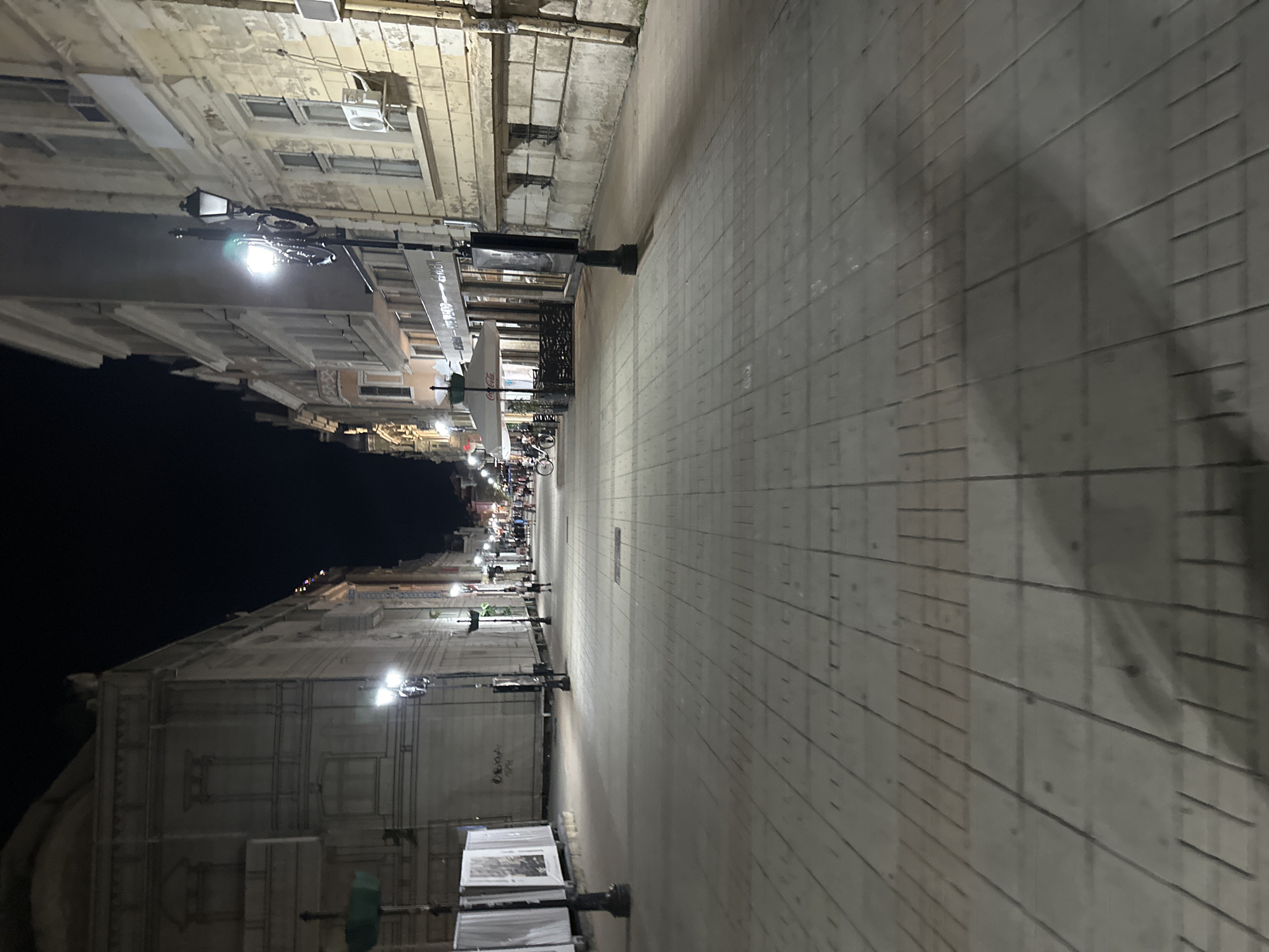 The image depicts a nighttime scene on a pedestrian street in Ruse, Bulgaria, likely the central pedestrian zone. The street is paved with light-colored square tiles, with darker rectangular tiles forming patterns along the length of the walkway. Tall, ornate streetlights with multiple lamps are spaced along the sides of the street, casting illumination onto the pavement. On the right side of the street, there are multi-story buildings with stone facades, windows, and some air conditioning units. A cafe with outdoor seating, indicated by umbrellas, is visible on the left side of the street further down. Several people are walking along the street, some in groups and some individually. There is also a bicycle parked on the left side of the street. The overall atmosphere is quiet and peaceful, with the artificial lighting creating long shadows on the ground.