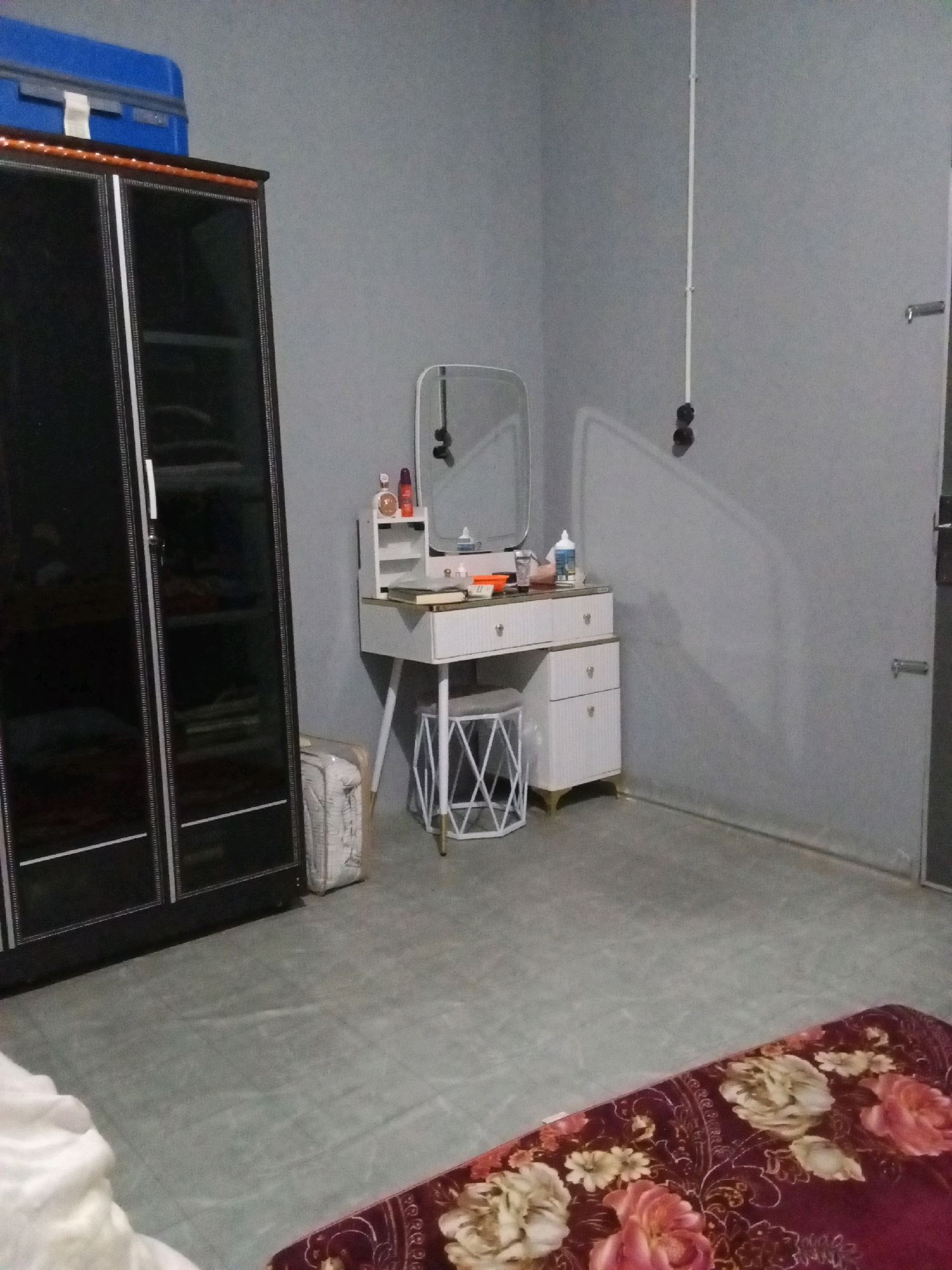 The image depicts an indoor scene, possibly a bedroom, with a modern aesthetic. On the left, a large, dark wardrobe with glass doors stands against a grey wall. A blue suitcase is placed on top of the wardrobe. To the right of the wardrobe, a white dressing table with a mirror and drawers is situated. Various cosmetic items and a book are arranged on the table. A white stool with a geometric design is placed in front of the dressing table. The floor is tiled in a greyish tone. In the foreground, a portion of a red blanket with a floral pattern is visible. The overall environment appears tidy and well-lit. No specific location in Gas Alam Badak I, Indonesia can be determined from the visual information. There are no people or discernible actions taking place in the image.