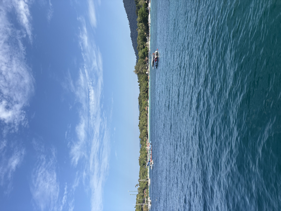 The scene depicts a clear-day panoramic view of the coastline of Ban Ko Li Pe, Thailand. The upper portion of the image shows a bright blue sky with scattered wispy white clouds. The lower two-thirds of the frame consists of calm, deep blue-green ocean water exhibiting minor surface ripples. The background features a white sandy beach fringed by dense tropical vegetation, including numerous palm trees and other foliage, which extends to a forested hillside on the right.
In the water, several vessels are visible:
* On the far left, multiple white-masted sailboats are anchored or moored near the shoreline.
* Closer to the center-left, a light-colored long-tail boat and another smaller vessel are present in the water near the beach.
* In the mid-right, a white and red trimaran-style speedboat, equipped with at least three outboard engines, is stationary on the water.
On the land, a tall antenna tower is discernible amongst the trees on the far left of the coastline. Several structures with reddish-brown roofs are partially visible through the vegetation along the beachfront. No individual persons or specific interactions are clearly identifiable within the scene.