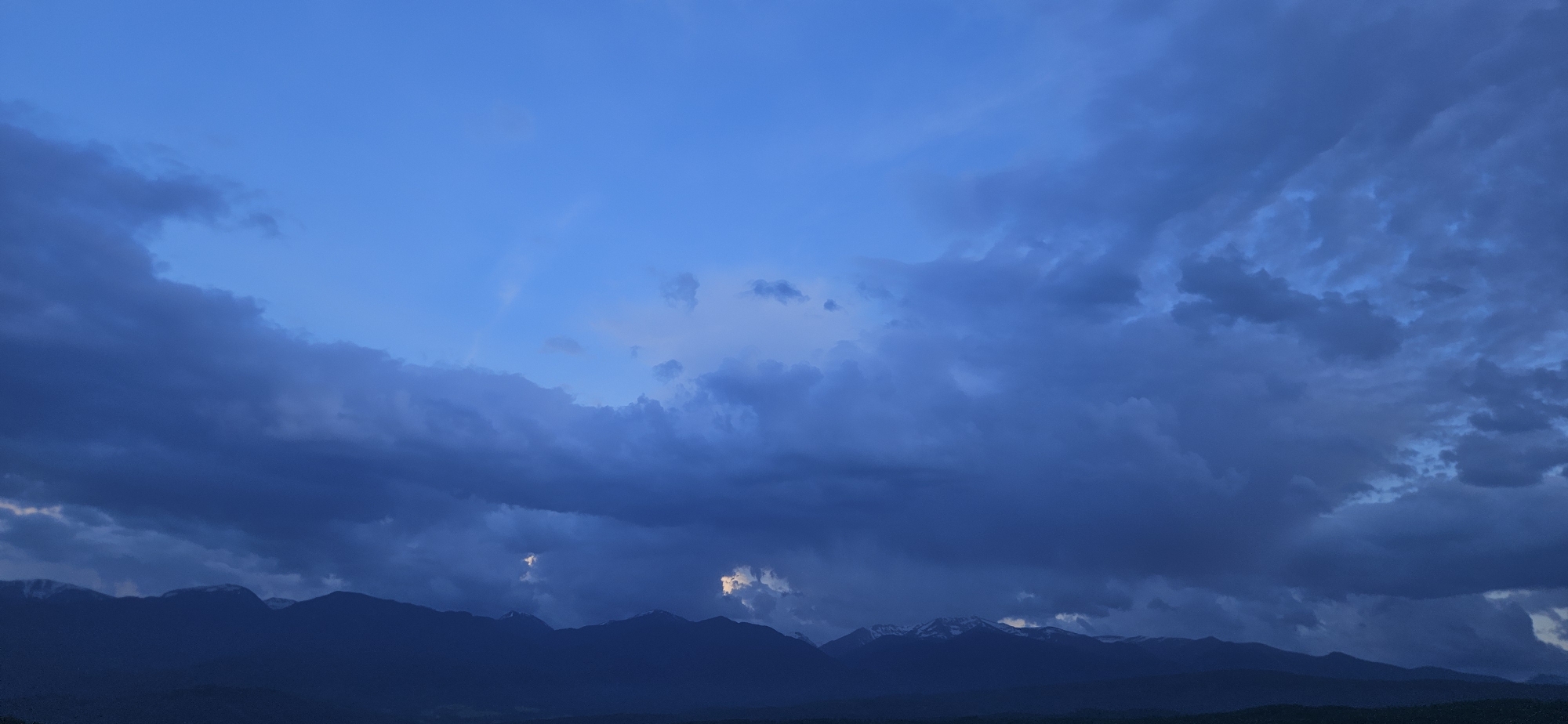 This image captures a dramatic and expansive sky over a mountain range in Gutsal, Bulgaria, likely at dusk or dawn. The dominant feature is the sky, a rich, deep blue that transitions from a lighter hue in the upper sections to a darker, more intense blue lower down. It is heavily overcast with a complex array of clouds.
Dark, voluminous storm clouds occupy the majority of the frame, particularly in the middle and lower sky, creating a sense of depth and intensity. These clouds exhibit various textures, from smooth, dense formations to more ragged and textured patterns, with some areas showing lighter reflections or patches where the light blue sky peeks through. Two distinct, brighter patches of light are visible near the mountain peaks, piercing through the heavy cloud cover.
Below the dynamic sky, a silhouette of mountains stretches across the horizon. These mountains are dark, almost black, against the lighter tones of the sky, emphasizing their vastness. Several peaks are discernible, some of which are capped with visible patches of snow, hinting at their elevation or the time of year. The land beneath the mountains is also dark and indistinct, suggesting forested terrain or valleys.
The overall mood is serene yet powerful, highlighting the grandeur of nature. There are no people, objects, or visible text in the image.