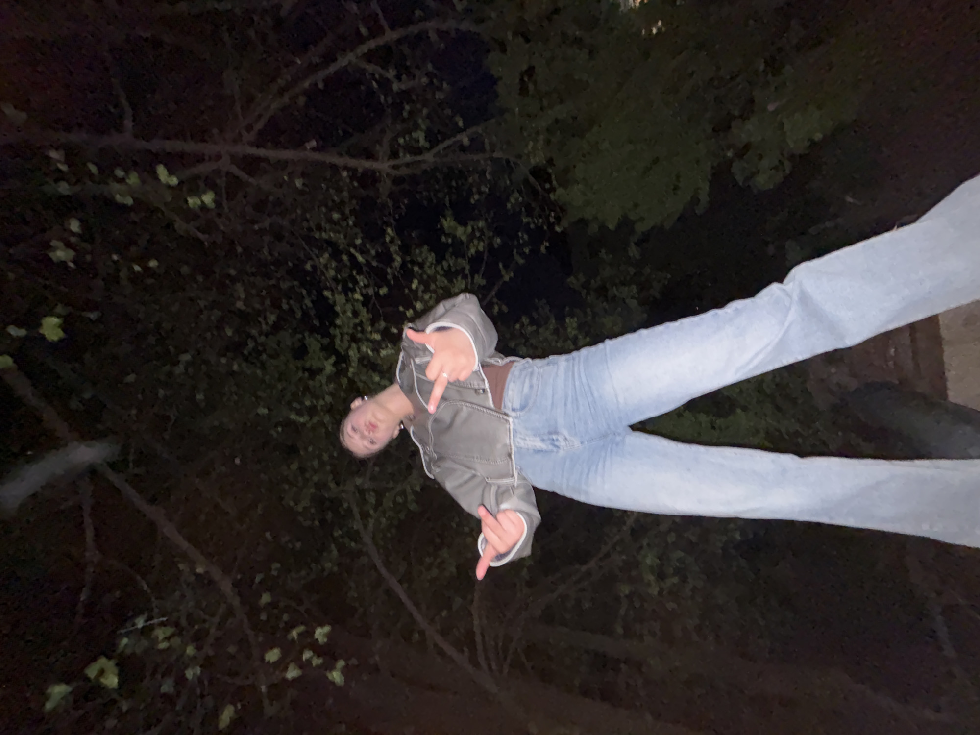 A young woman stands outdoors at night. She is wearing jeans, a brown top, and a gray jacket, and is extending her middle finger with both hands. The background is dark, with trees and foliage visible. It is not possible to verify the location in the city of Фоки, Russia based on the visual information provided.