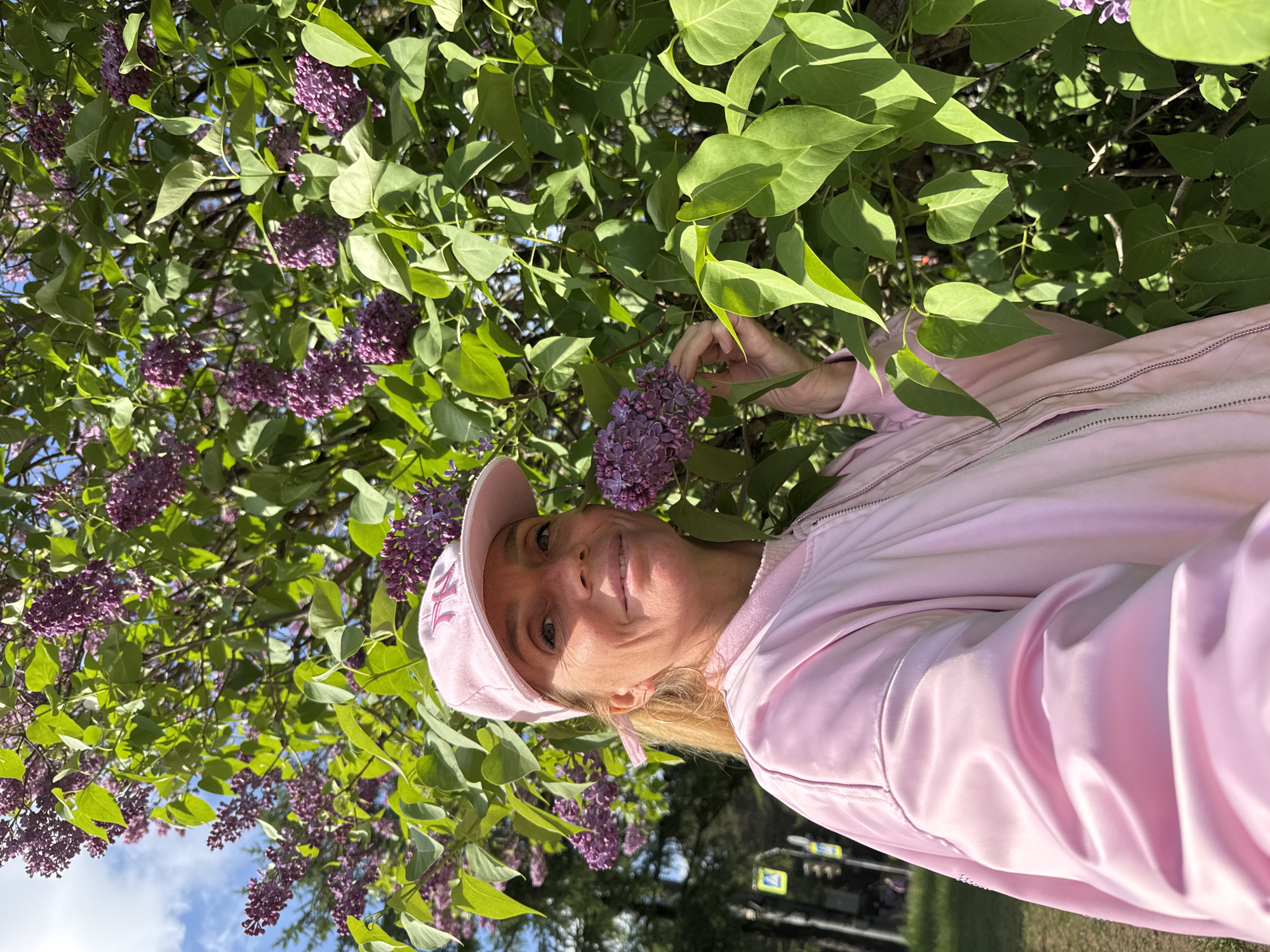 The image captures a woman in front of a lilac bush, likely in Saint Petersburg, Russia, based on the style of street signs visible in the background. The woman, wearing a light pink baseball cap and a matching jacket, is holding a cluster of lilac blossoms. The background includes a dense lilac bush with both green leaves and purple flowers, as well as a partial view of a cloudy sky and some street furniture.