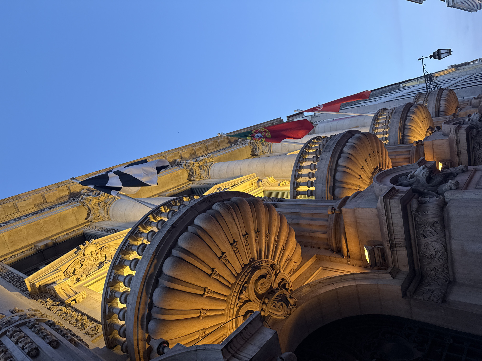 The image shows the exterior of a grand building in Lisbon, Portugal, at dusk. The building features ornate architecture with classical columns, decorative balconies, and intricate stonework. Several flags are visible, including the Portuguese flag and a black and white checkered flag. The building is illuminated by warm, artificial lights, highlighting the architectural details. A lamppost is also visible, adding to the old-world charm of the scene. The sky is a clear, deep blue, indicating the transition from day to night. The perspective is from a low angle, looking up at the building, emphasizing its grandeur.