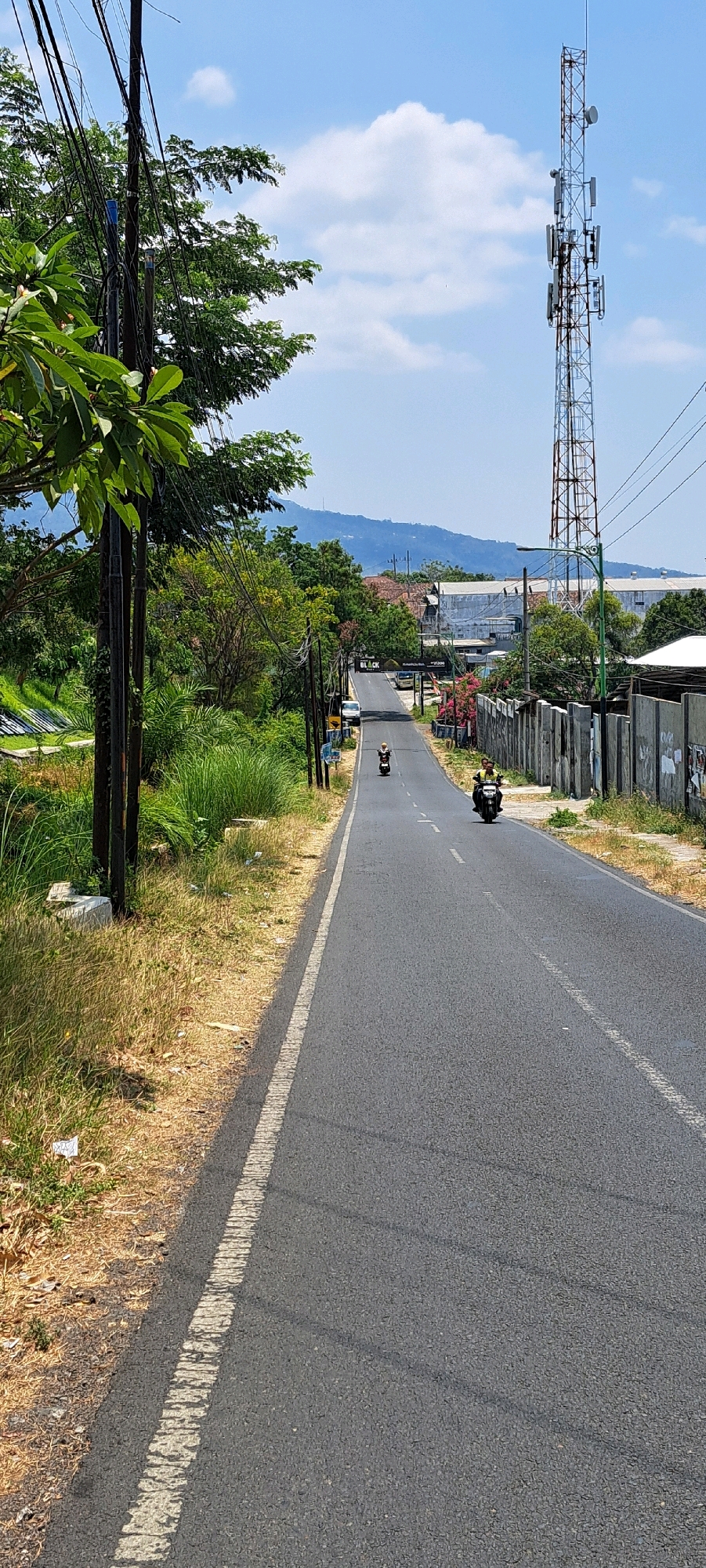 This is a medium-shot, vertical, eye-level, outdoor, daytime photograph showing a paved road in Pandaan, Indonesia. The road curves slightly and heads into the distance, where it meets a backdrop of rolling hills under a bright blue sky with scattered white clouds.
On the left side of the road, there is a utility pole adorned with numerous black wires, and lush green foliage. The ground beside the road is covered in dry, yellowed grass with some scattered litter.
On the right side of the road, a gray concrete fence runs along the edge, with some posters affixed to it. Beyond the fence, various buildings and structures are visible, including a prominent telecommunications tower with multiple antennas.
Two individuals on motorcycles are present on the road. The first motorcycle, closer to the foreground on the right, is ridden by a person wearing a yellow shirt and a dark helmet. Further up the road, another motorcycle is visible with a person wearing a light-colored shirt and a dark helmet.
The overall scene depicts a semi-rural or suburban environment, typical of the outskirts of a town like Pandaan, with a blend of natural elements, infrastructure, and human activity.