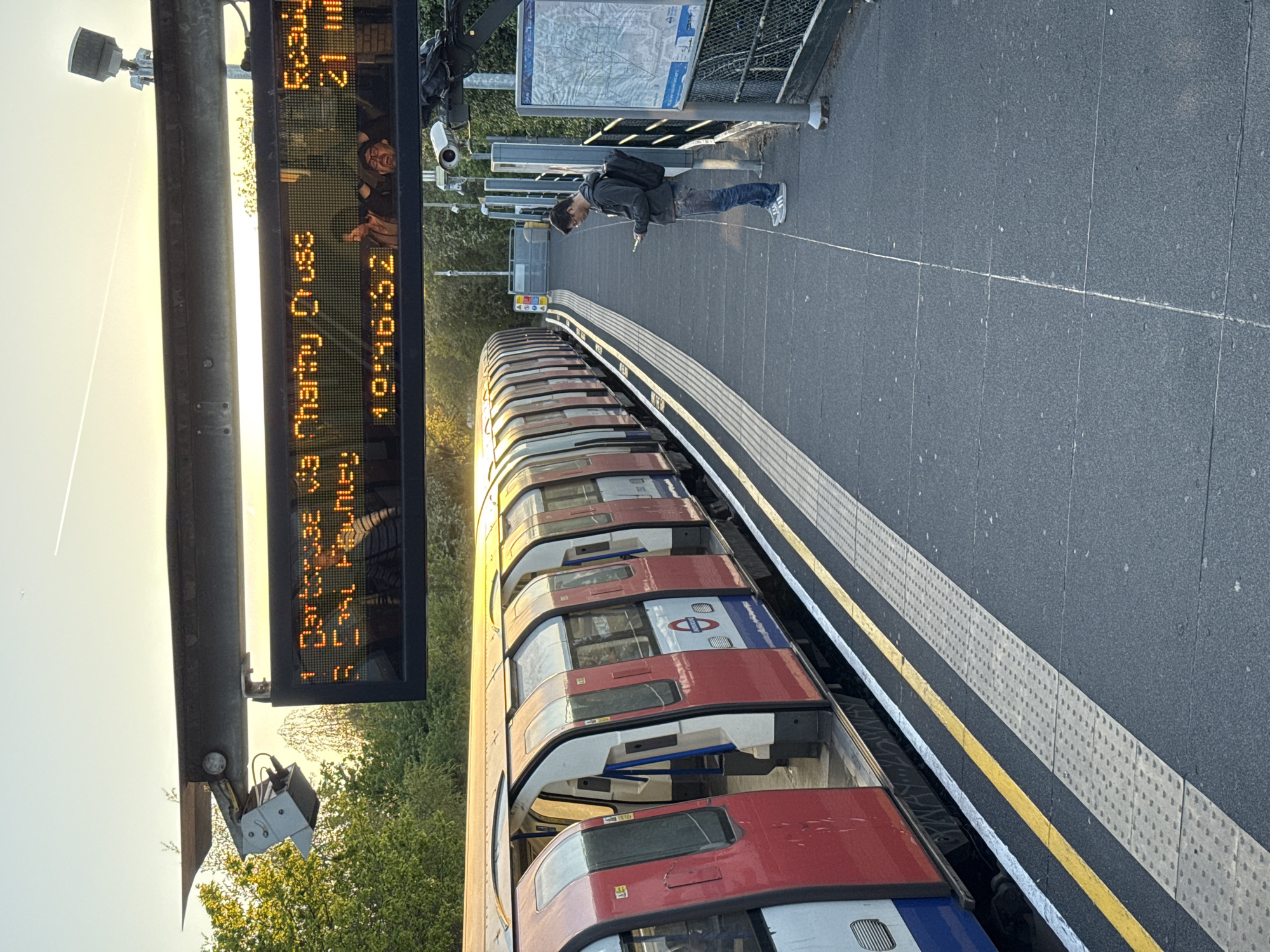 An outdoor, eye-level shot depicts a London Underground train at a station. A person stands on the platform looking at their phone. An electronic sign displays train arrival times, showing "1 Dartford via Charing Cross...19:46:52" and "Roadway...21 min." The sky indicates either sunrise or sunset. The environment includes trees and station signage.