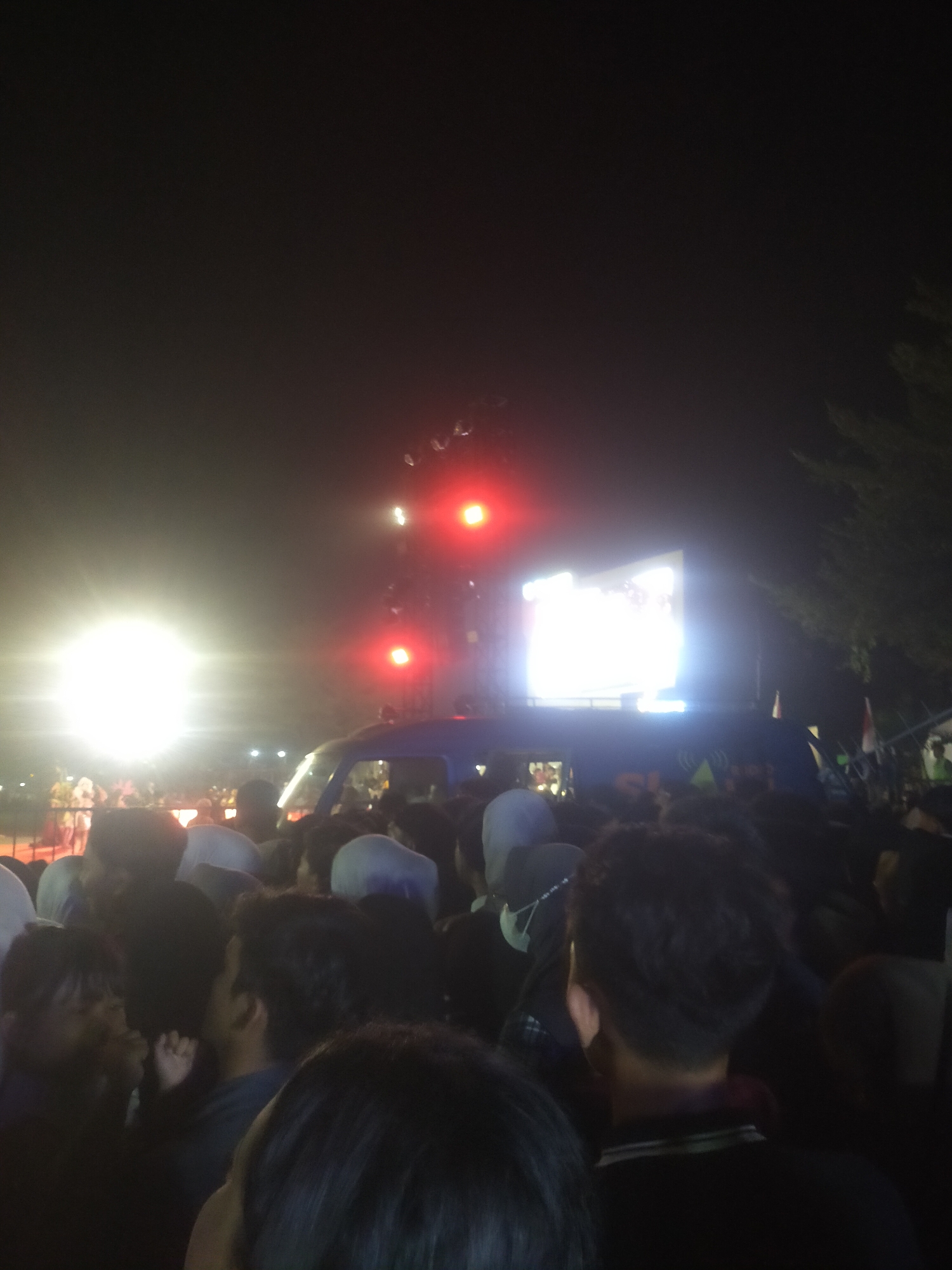 The image shows a crowd of people gathered outdoors at night, presumably at an event in the city of Slawi, Indonesia. A large, brightly lit screen is visible in the background, along with stage lighting, including red lights. A dark-colored vehicle, possibly a van or bus, is positioned among the crowd and in front of the screen. Many individuals are facing towards the screen and lights, suggesting they are watching a performance, concert, or screening. The scene is illuminated by artificial lights, creating a somewhat hazy atmosphere. The overall impression is of a lively public gathering.