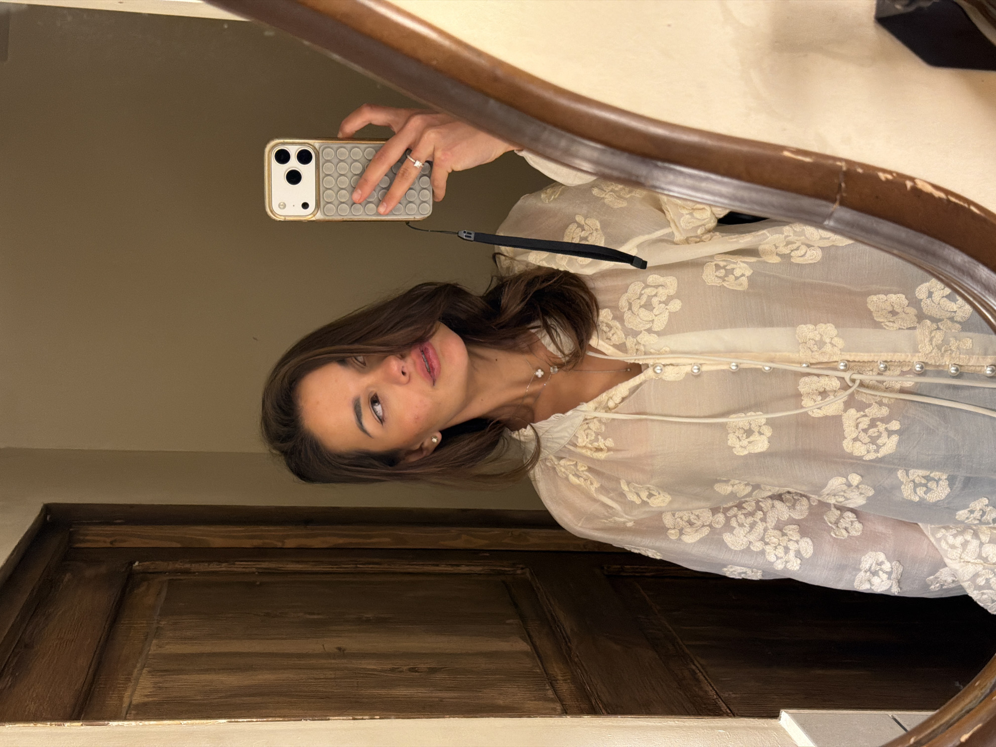 A woman is captured in a mirror selfie, showing her from the upper chest to just below the waist. She has medium-brown hair, dark eyes, and fair skin, with her head tilted and her gaze directed to her right (viewer's left), her tongue slightly visible between her lips. She wears small stud earrings and a layered necklace featuring a pearl drop and clover-shaped pendants. Her attire consists of a sheer, light cream or off-white blouse adorned with embroidered floral patterns, a V-neckline, and a front placket secured by small pearl buttons, with a tie detail at the collar. She holds a gold-framed smartphone, identifiable as an iPhone with a triple-lens camera system, encased in a light grey silicone "pop-it" style case. A black wrist strap is attached to the phone, which she holds with her left hand, revealing a ring with a clear stone on her left ring finger. The background reflects a warm-toned wall and a dark brown wooden door with vertical paneling on the left. The mirror's curved, dark wooden frame shows signs of wear, including chipped finish. The scene is located in a room in Tbilisi, Georgia.