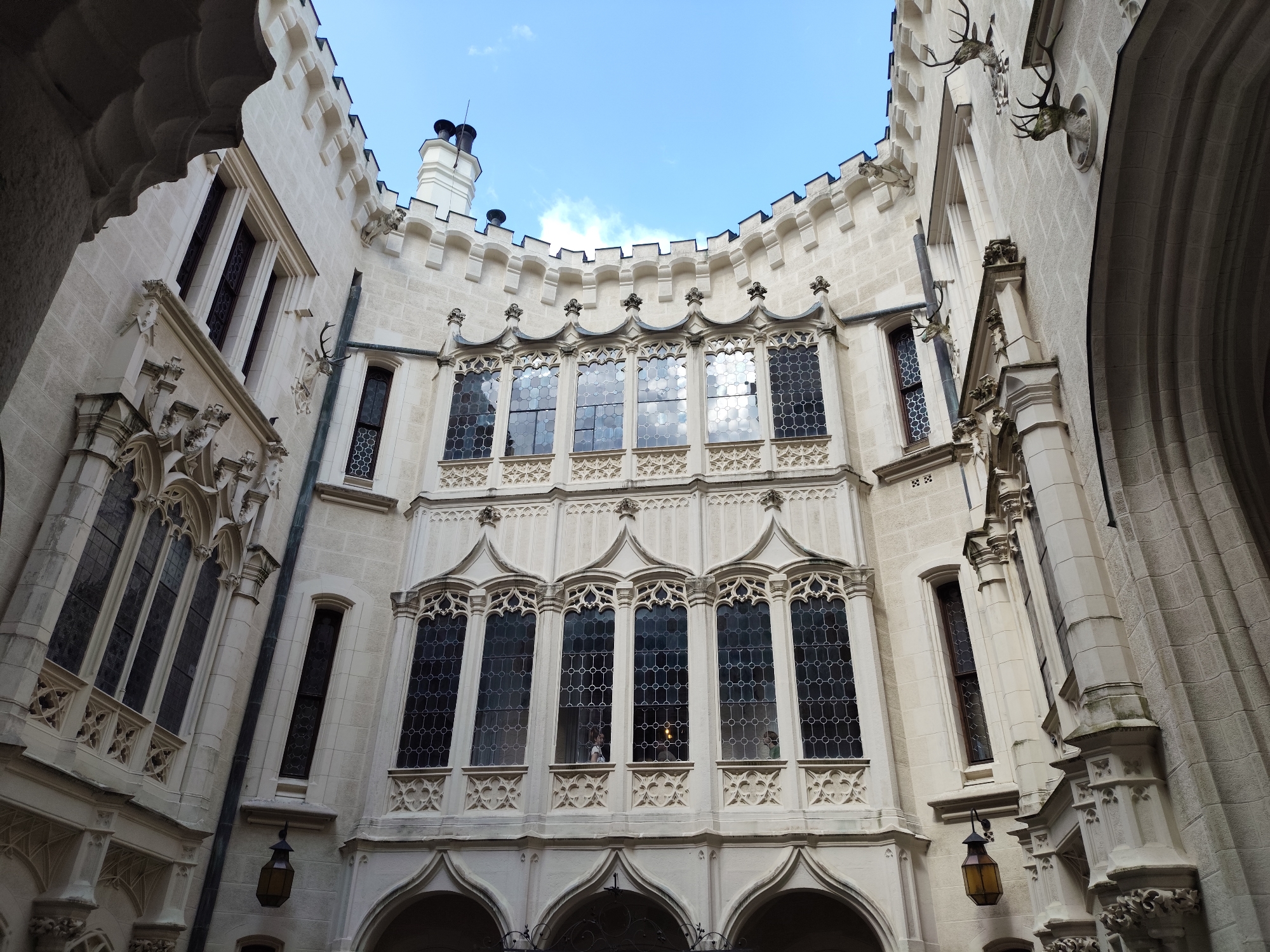 This is a low-angle, wide shot of the interior courtyard of Hluboká Castle, located in Hluboká nad Vltavou, Czechia. The castle is constructed of light-colored stone and features intricate Gothic Revival architecture. The courtyard walls rise up on all sides, creating a sense of enclosure. On the central facade, a series of tall, narrow windows with decorative tracery are visible. Above these windows, a crenellated parapet runs along the top of the building. On the outer walls, more windows and architectural details, including gargoyles and what appear to be mounted deer antlers, can be seen. The sky above is a clear blue with a few scattered white clouds. Hanging lanterns with amber glass are attached to the walls of the courtyard. The overall impression is one of grandeur and historical significance.