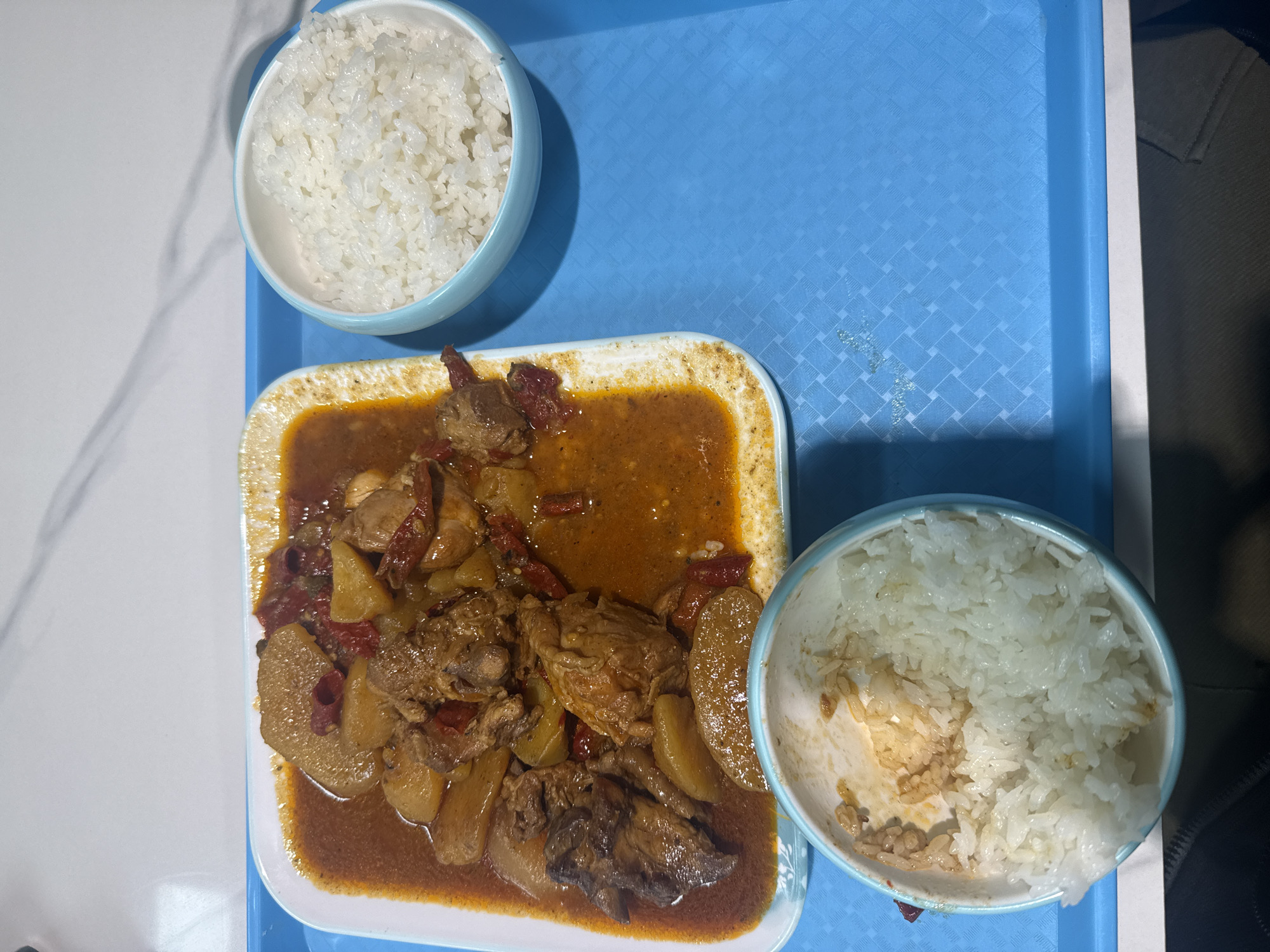The image shows a meal on a blue tray. There is a square white plate with chicken and potatoes in a reddish-brown sauce. Two small bowls of white rice accompany the plate, one nearly full and the other partially eaten. The tray rests on a white surface. The image provides no information to confirm the location as Yuzhong District, China.