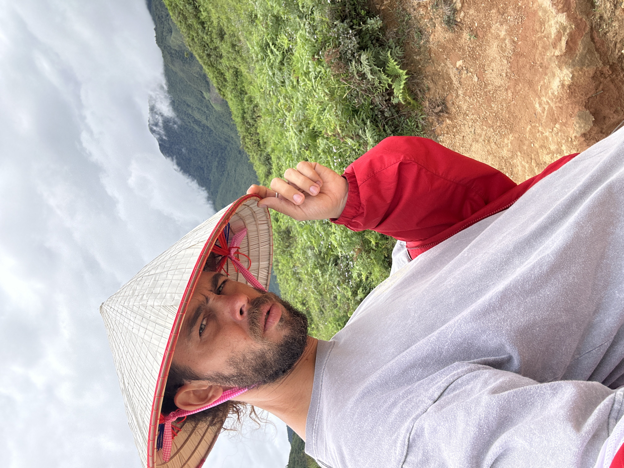 A man wearing a conical hat and a gray long-sleeved shirt is captured in a selfie outdoors. He is positioned on the left side of the frame, with his body angled towards the camera and his gaze directed at the lens. His right hand is raised, lightly touching the brim of his hat, and he is wearing a red jacket over his shirt. The background reveals a lush green mountainside covered in dense vegetation, with a cloudy sky overhead. The ground in the foreground appears to be a reddish-brown dirt path. The scene is set in the mountainous region of Ta Xua, Vietnam.