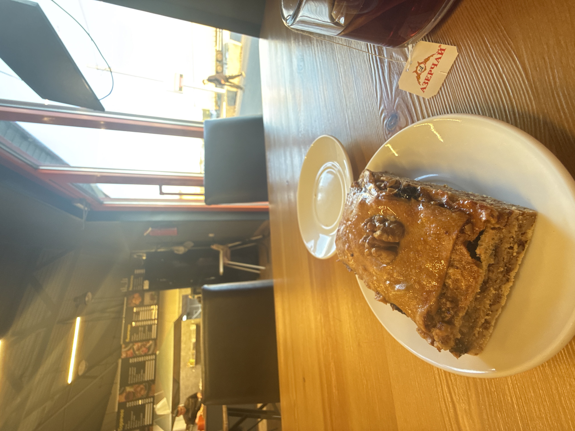 A slice of cake on a white plate and a cup of tea with the "Azerchai" tea bag on a wooden table are located inside a restaurant in the city of Unknown, Unknown. A chair, window, and menu can be seen in the background. The photo is taken at an oblique angle.