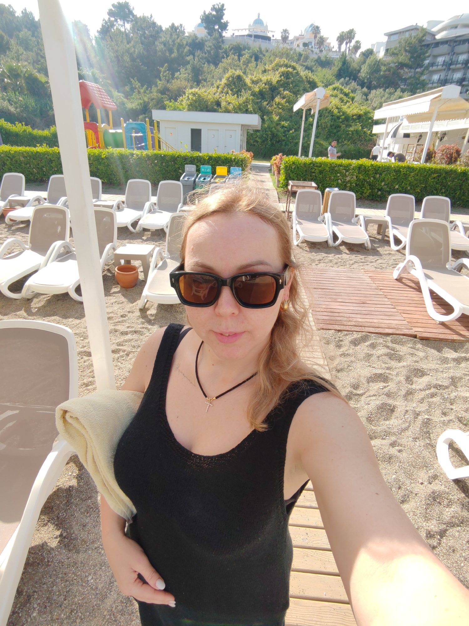 A woman stands on a sandy beach and wooden decking, amidst rows of empty deck chairs. The woman wears a black tank top and large sunglasses. She holds a light-colored towel in her left arm. Behind her is a beach scene with a playground, a small white building, and several sun shelters. In the background, a hillside is covered with dense green trees, and architectural structures with domes are visible. Based on these architectural details and the presence of a beach resort, the location is likely the city of Alanya, Turkey.