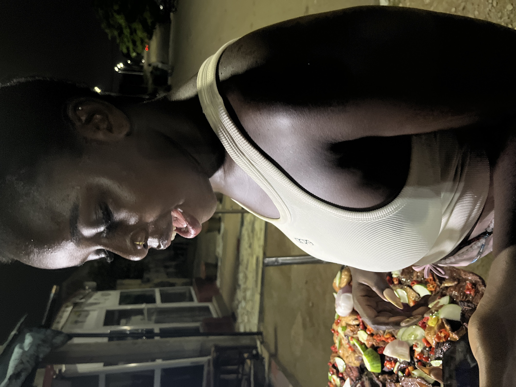 A young Black woman is pictured at night in an outdoor setting, likely in Abuja, Nigeria, possibly at a street food stall or market. She is wearing a light-colored, ribbed tank top and has a nose ring. Her mouth is open as if she is speaking or about to eat. In front of her is a large platter of food, which appears to be a variety of meats, vegetables such as cucumbers and onions, and chili peppers. Her right hand is reaching towards the platter, suggesting she is about to serve herself or has been preparing the food. The background is dark, with some indistinct structures and streetlights visible, indicating an urban environment at night. The overall lighting suggests the scene is illuminated by artificial light sources.