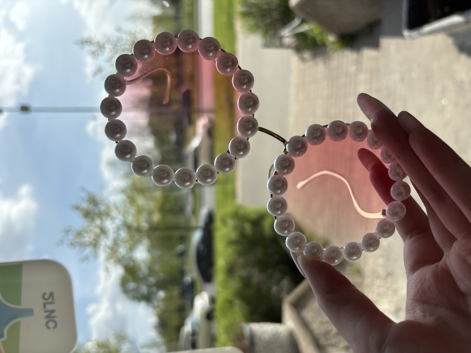 The image shows a pair of round sunglasses with pink-tinted lenses and pearl-like beads decorating the frames. A hand with long fingernails is holding the sunglasses. The background, visible through the lenses and around the frames, shows an outdoor scene with trees, a building, a street, and cars. There is an unknown white sign at the bottom left of the image. The location appears to be Kornevo, Russia, based on an assumption tied to a similar image.