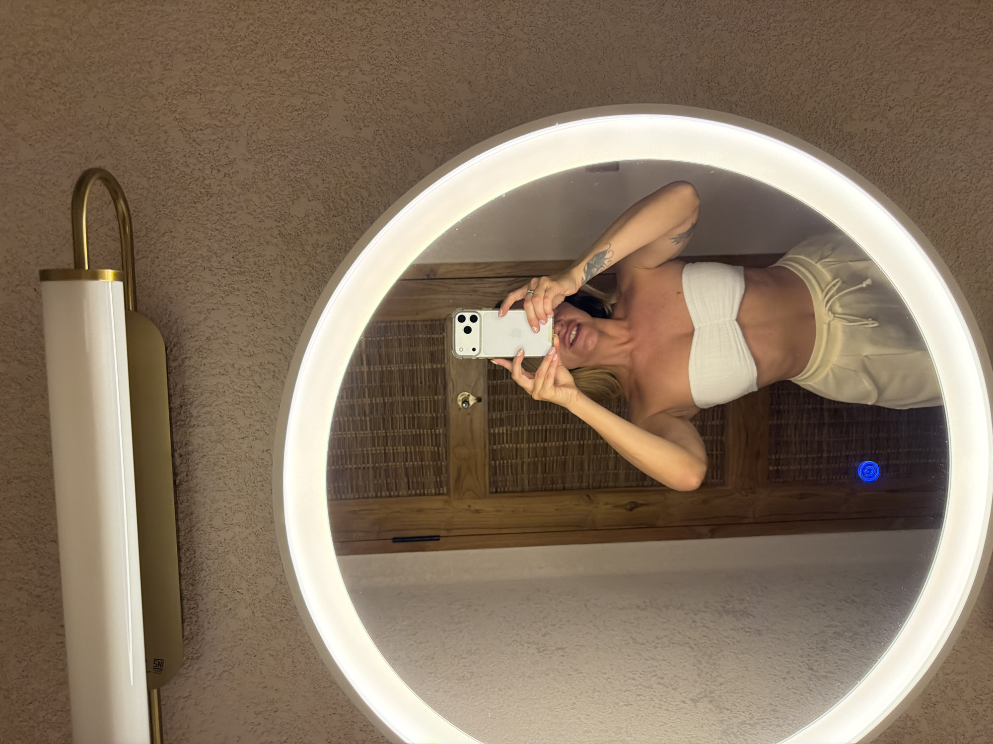 An indoor, vertically oriented mirror selfie captures a woman taking a picture with her smartphone in what appears to be a bathroom or changing area in Mengwi, Indonesia. The woman is reflected in a large, circular mirror that is brightly backlit with an integrated LED light strip, creating a glowing halo effect. She holds a light-colored smartphone with a clear case, covering most of her face but revealing a smiling mouth. Her left arm, adorned with a tattoo, is visible as she holds the phone. She wears a white, textured bandeau top and light-colored (possibly cream or pale yellow) drawstring loungewear pants. A ring is visible on a finger of her left hand.
To the left of the mirror, a modern wall sconce with a gold-colored metallic frame and a long, white cylindrical shade is mounted on a textured, light brown or beige wall. A small "SNI" mark is visible on the side of the sconce. Inside the mirror's reflection, behind the woman, elements of the room's decor are visible, including a wooden structure and a woven material, possibly part of a cabinet or wall covering, suggesting a natural and resort-like aesthetic. A small, blue illuminated circle, likely a touch control for the mirror's light, is visible on the mirror's surface in the lower right quadrant. The scene is well-lit by the mirror's illumination and the sconce, suggesting an indoor setting where artificial light is primary.
