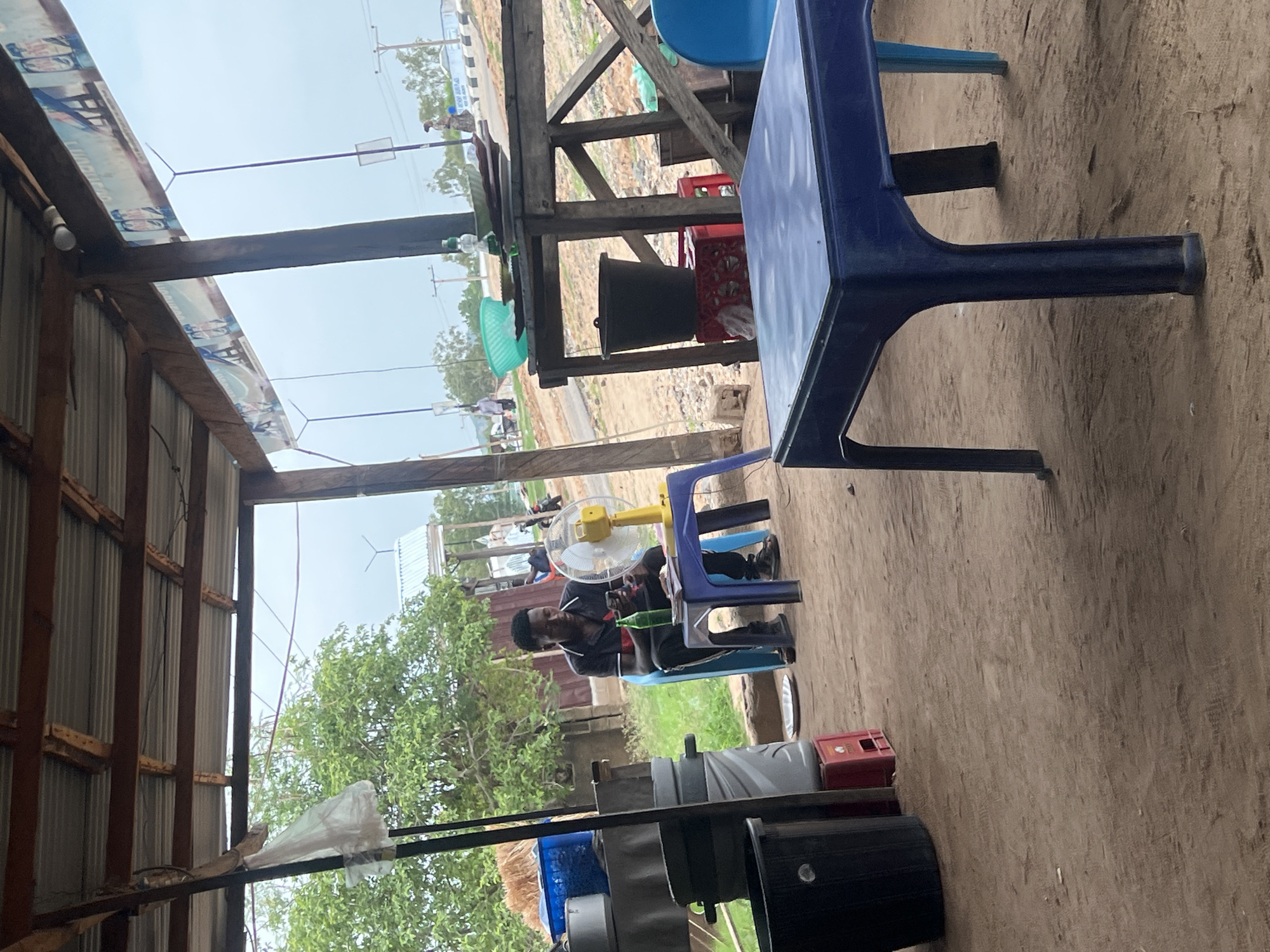 The image depicts an outdoor scene in Jalingo, Nigeria. An individual is seated at a plastic table under a makeshift shelter with a corrugated metal roof. A small electric fan is positioned near the person. The surrounding environment consists of sandy ground, plastic chairs, containers and a road is visible in the background.