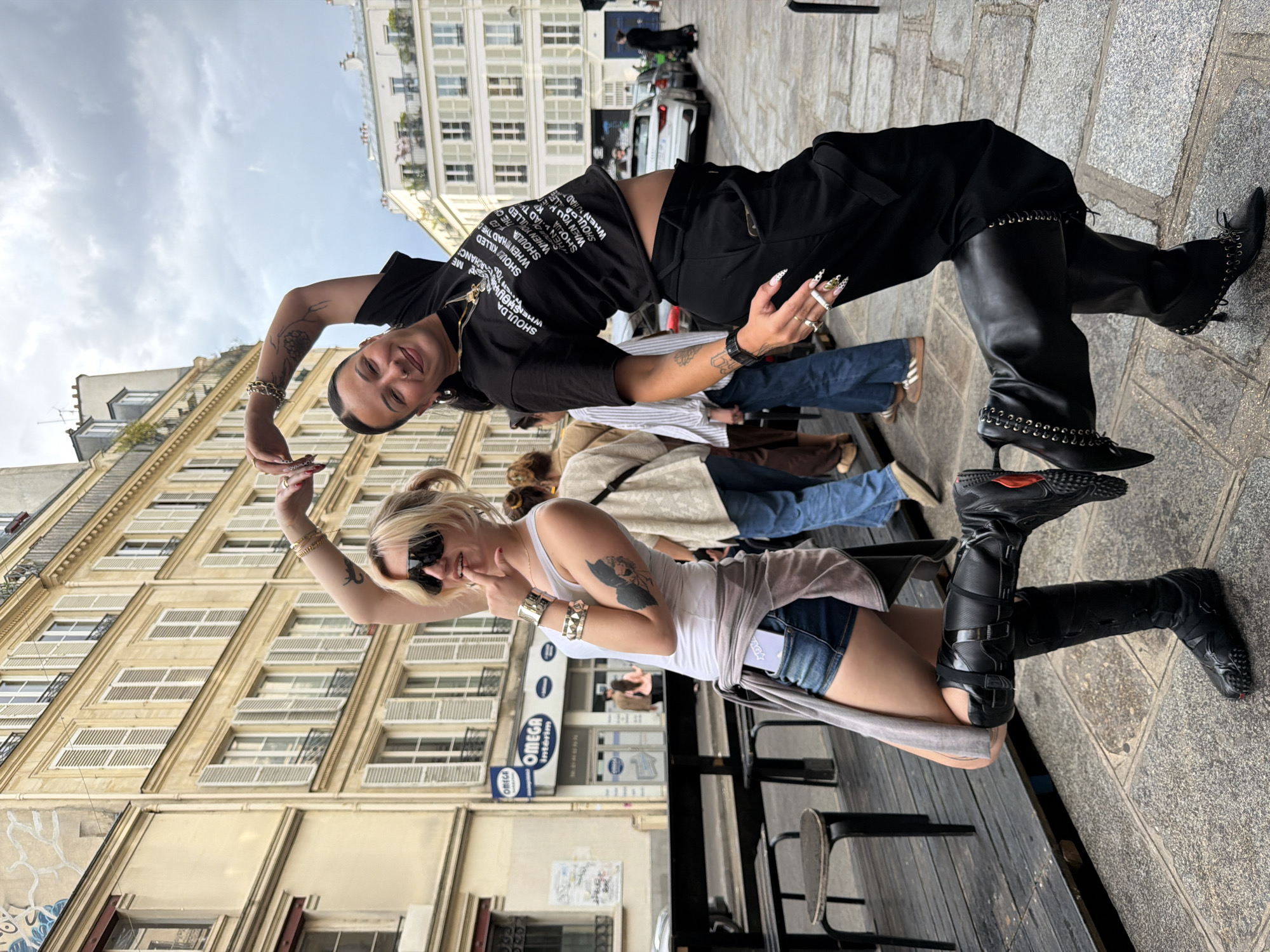 Two young women pose outdoors in Paris, France. The woman in the foreground wears a white tank top, denim shorts, black boots, and sunglasses, with a gray sweater wrapped around her waist. The woman behind her wears a black top with white text, black pants, and black boots with metal embellishments. Both women have tattoos on their arms and are making a heart shape with their hands. Behind them is a building with multiple windows. Other pedestrians are visible in the background, along with a parked white car. An "OMEGA Intérim" sign is visible on a building.