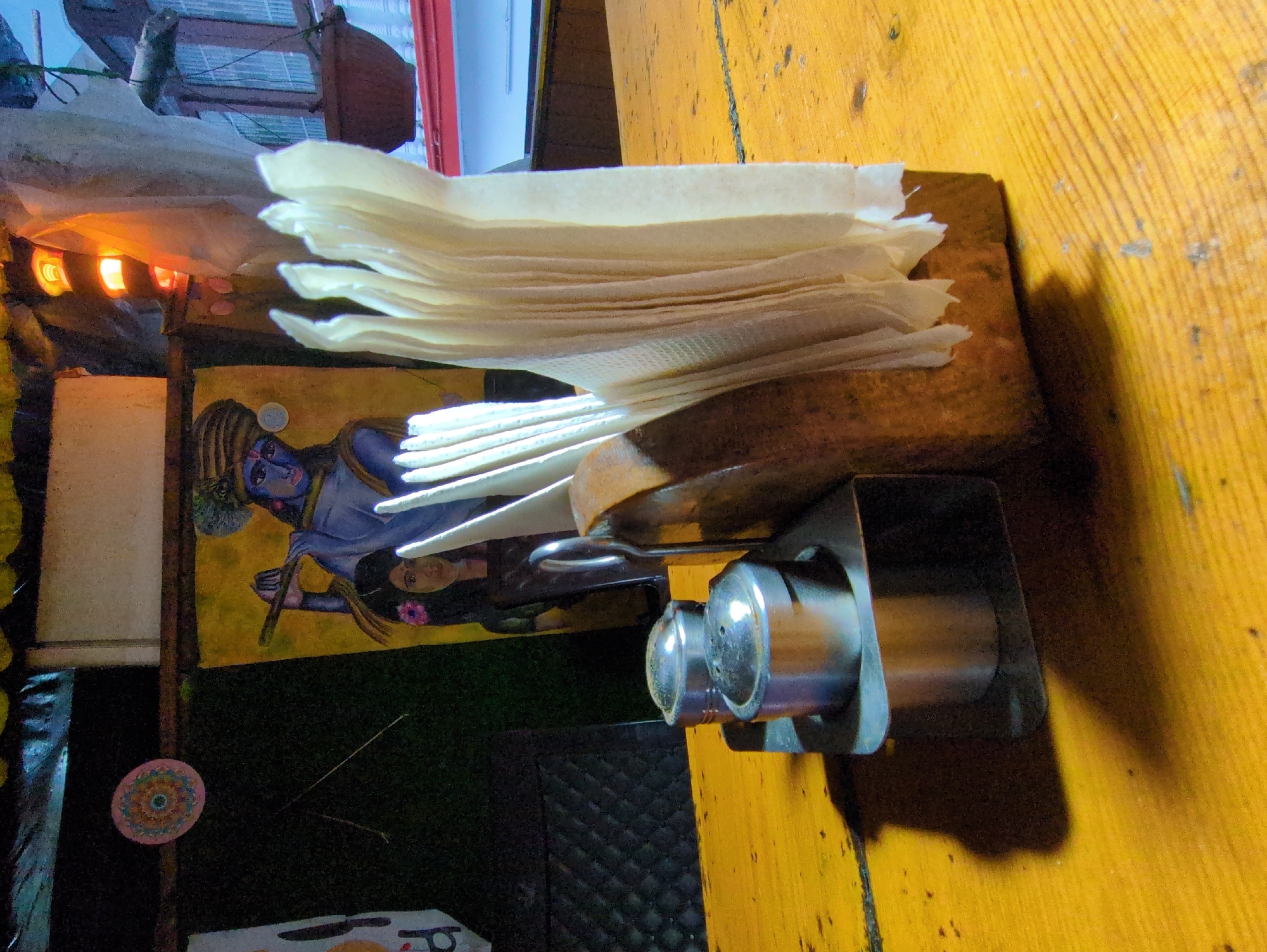 In Bhagsu, India, this image captures a close-up of a yellow wooden table, likely in an outdoor or semi-outdoor setting, perhaps a cafe or restaurant. The focus is on a rustic wooden napkin holder filled with white paper napkins, positioned next to a metal salt and pepper shaker set. Behind these items, a colorful painting hangs on a dark background. The painting depicts a figure, possibly a deity like Krishna, playing a flute, with a smaller figure to the side. The overall ambiance suggests a casual, possibly bohemian, environment.