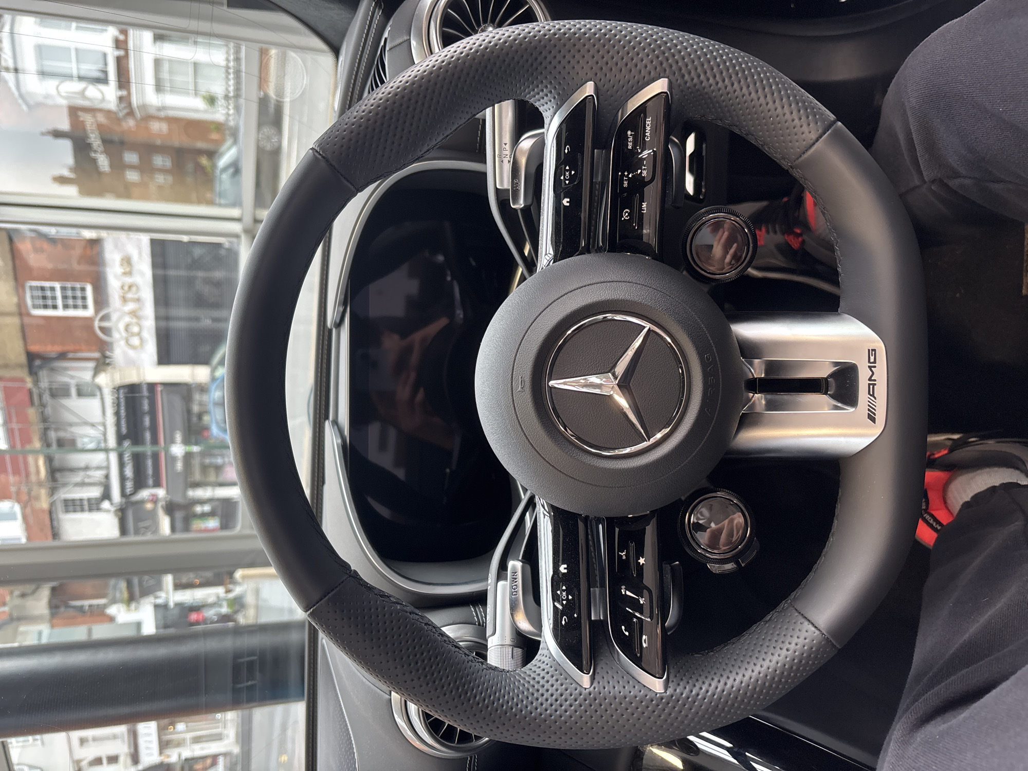 The image depicts the steering wheel of a Mercedes-AMG vehicle, prominently featuring the Mercedes-Benz logo at its center and "AMG" branding on the right spoke. The steering wheel is black leather with a perforated grip. Various control buttons and dials are integrated into the wheel's design.
Through the car's window, the reflection reveals a building, with the word "COATS" partially visible on a sign, potentially indicating a clothing store. The architectural style suggests a location in London, United Kingdom. Also, there is a partial view of a person wearing jeans sitting in the driver seat.