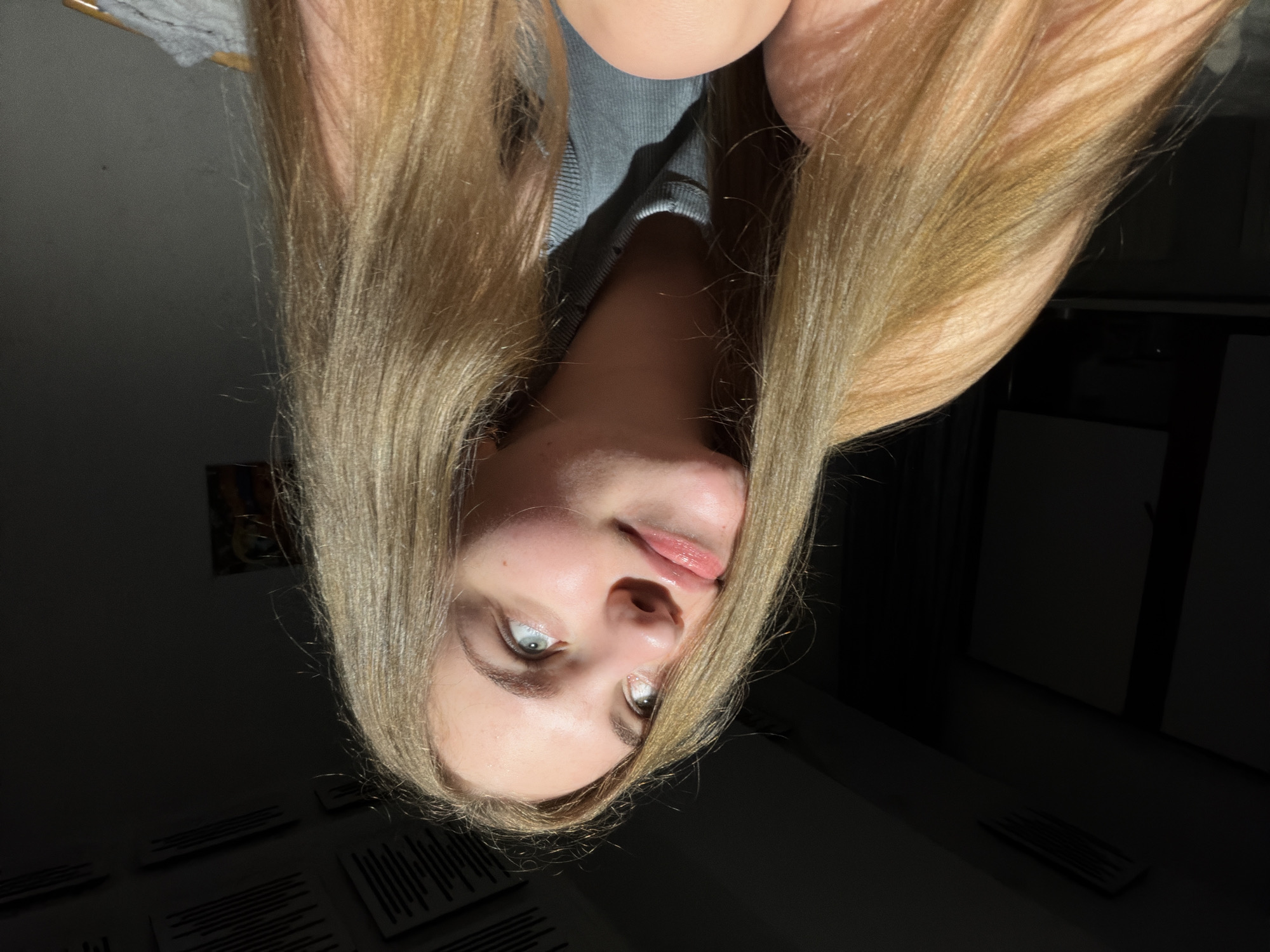 A young woman with long, blonde hair is captured in a low-angle, indoor shot. She is looking upwards and to her left, with her eyes slightly narrowed and her lips parted. Sunlight illuminates her face from the left, creating a dramatic contrast with the dark background. She is wearing a gray ribbed tank top. The environment appears to be a room with dark walls and a ceiling with visible acoustic tiles. There is a hint of a picture on the wall in the background. The scene is devoid of any identifiable city landmarks or context that would place it specifically in Dnipro, Ukraine.