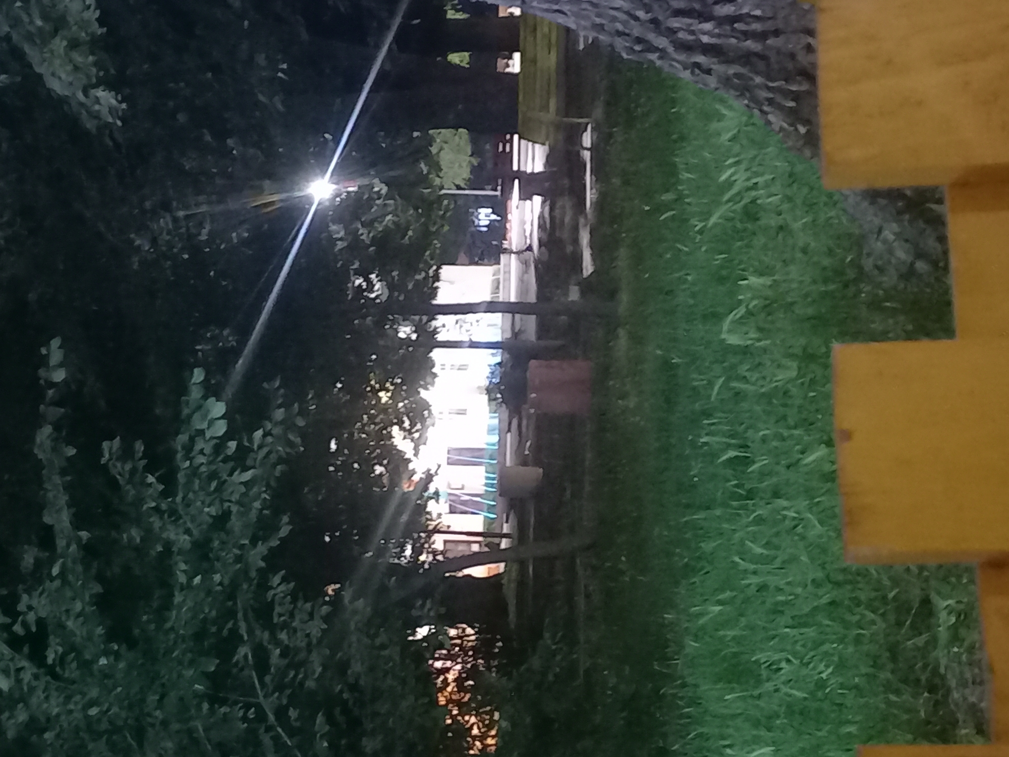 The image depicts an outdoor night scene in Parvomay, Bulgaria, likely a public green space or park. In the foreground, a low, yellow-orange wooden fence or barrier partially obstructs the view from the bottom of the frame. Beyond the fence, a patch of green grass extends into the midground.
The midground is dominated by several trees with dense, dark green foliage, which occupy the upper sections of the image. A bright artificial light source, positioned in the upper-right quadrant, emits a strong, star-shaped lens flare that diagonally traverses the scene. Beneath the trees, park amenities are visible, including a light-colored bench and what appears to be a dark table or another bench. A prominent tree trunk is partially visible on the far right, adjacent to the foreground fence. Towards the left-center, a light brown cylindrical object, possibly a planter or rubbish bin, stands on the ground. A reddish-brown rectangular object is situated on the ground on the far left.
In the background, through gaps in the tree canopy, several illuminated buildings are discernible. These structures are predominantly light-colored (white or off-white) and feature dark rectangular openings, likely windows or doorways. Areas on the left side of the background buildings exhibit a distinct blue light, suggesting either reflective surfaces or decorative lighting. No individuals or discernible actions are present in the scene.