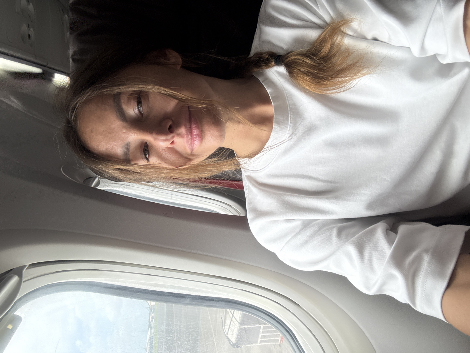 A woman is seen from an unconventional angle, possibly a selfie, seated next to an airplane window. She is wearing a white, long-sleeved t-shirt, and her hair, which appears to be brown, is pulled back in a loose braid. The window next to her reveals a view of an airport, presumably in Kuta, Indonesia, evidenced by the tarmac and potentially identifiable architectural features. The angle suggests she may be reclining or in a slightly tilted position.