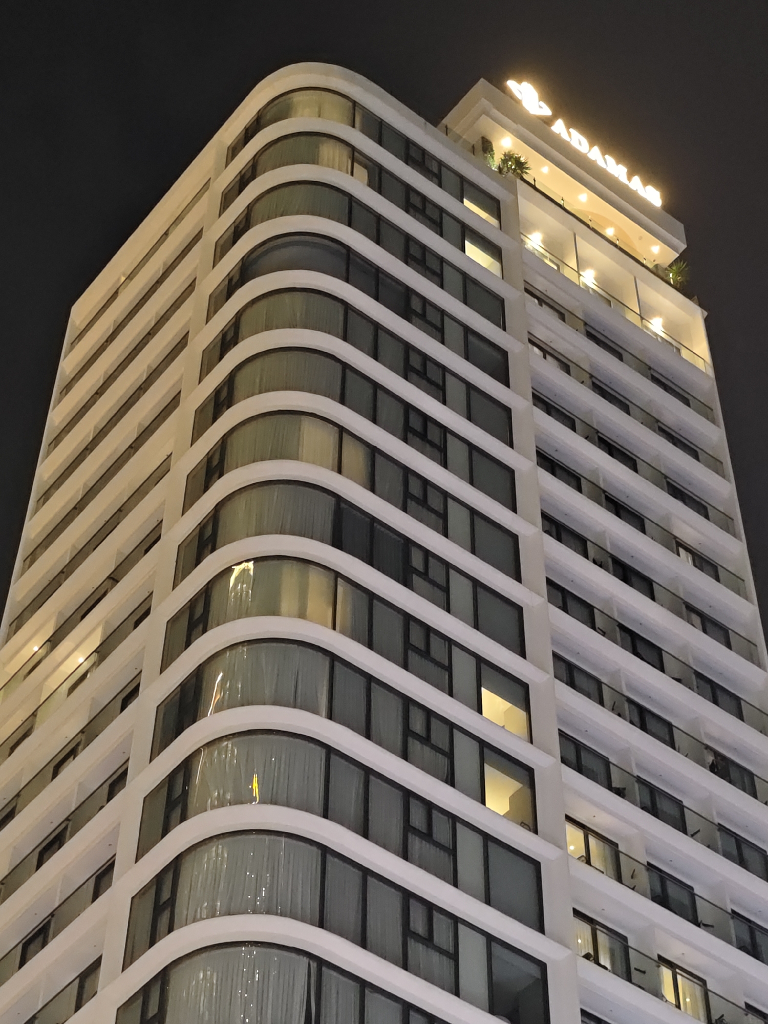 This media file features a nighttime exterior shot of a modern, high-rise building in Nha Trang Ward, Vietnam. The building, likely a hotel or apartment complex, has a distinctive curved facade with white trim separating floors. Numerous windows, some with curtains visible, line the structure, and many are illuminated from within, suggesting occupancy. Balconies with glass railings are also present on several levels.
At the very top of the building, illuminated signage reads "ADAMAS," with a stylized logo above it. This signage and some decorative lighting on the uppermost terrace indicate that the building is likely a commercial establishment. The overall impression is one of urban sophistication and nighttime activity. The dark sky, devoid of stars or moon, suggests a potentially overcast night or late evening hours. The lighting from the building casts a warm glow against the dark background.