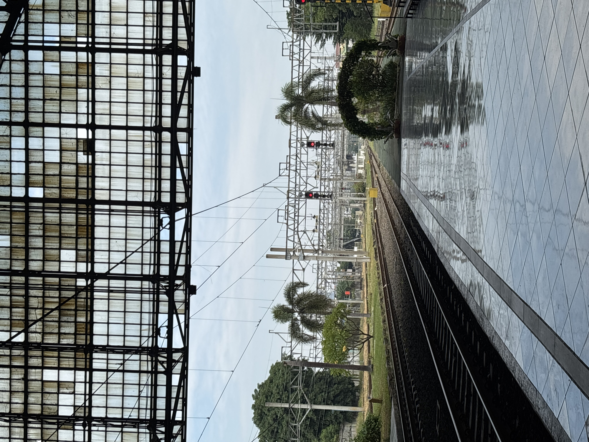 This image captures a train station environment, likely on an overcast day, with a focus on the tracks, platform, and overhead structures. The scene is viewed from a platform, looking down the railway line.
On the left side, a large, dark, grid-patterned roof structure, appearing to be made of glass or translucent panels, extends overhead. This canopy provides shelter, and its panels show signs of weathering or dirt.
In the mid-ground, multiple railway tracks run horizontally from the foreground into the distance. Above the tracks, a dense network of silver-colored overhead electrical lines (catenary system) is supported by numerous metallic gantries and poles, suggesting an electrified railway. Several red signal lights are visible along the tracks, indicating a stop. Scattered among the tracks and in the background are various trees, including several palm trees, and other lush green vegetation. A prominent, dark green, circular shrub or topiary is situated near the right side of the tracks.
On the right side, a wide, light-colored platform made of square tiles arranged in a diamond pattern extends towards the horizon. The platform surface appears wet and highly reflective, mirroring the overhead structures, trees, and parts of the sky in distorted patterns. The sky itself is bright but uniformly gray, suggesting an overcast day.
There are no people visible in the image, and no specific activities are taking place. The wet platform hints at recent rain. The overall impression is one of a quiet, somewhat industrial yet green, transit hub. No visible text is present in the image. The presence of palm trees and the general climate implied by the vegetation align with the provided context of North Jakarta, Indonesia.
