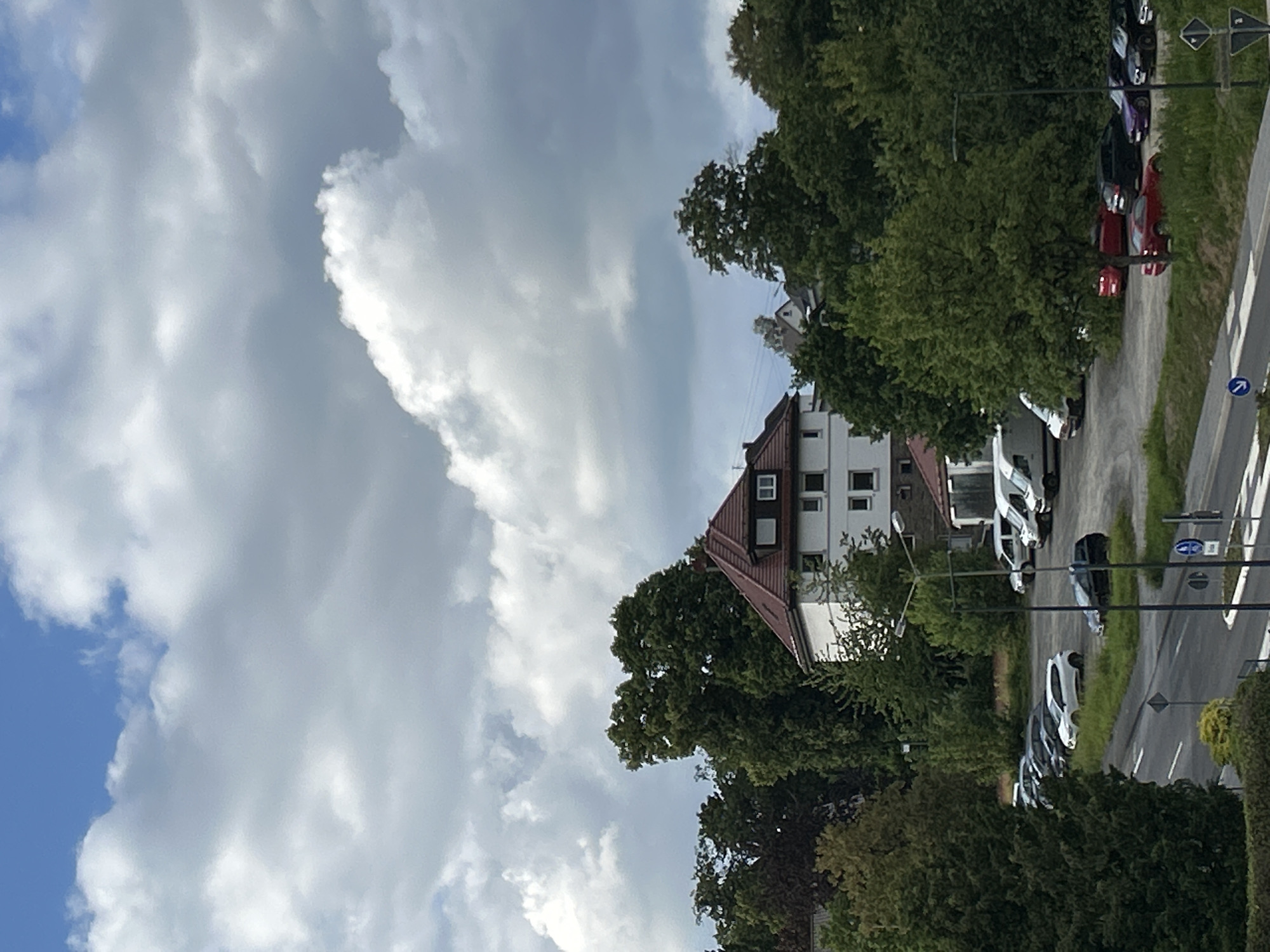 In Waldbröl, Germany, a residential building with a red tiled roof is visible amidst a dense green canopy of trees. In front of the building, a parking lot contains several vehicles, including white vans, a white hatchback, and a dark sedan. Adjacent to the parking lot is a road with white lane markings, where a dark sedan is parked. The sky above is filled with dramatic, billowing clouds against a blue backdrop.