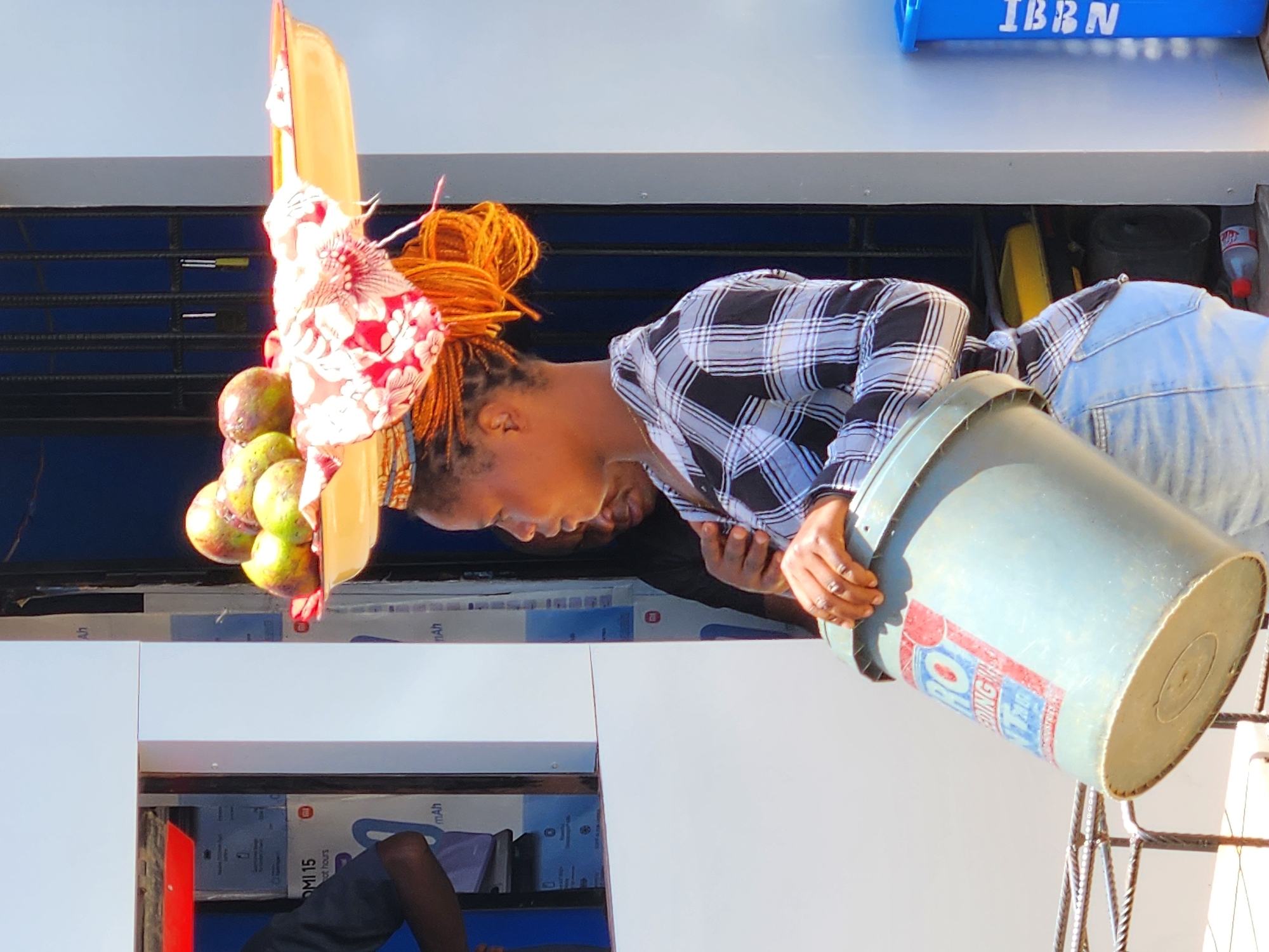 A woman with orange hair tied up is leaning over a white counter, supporting a bucket. She is wearing a blue and white plaid shirt and light blue jeans. On her head, she carries a yellow tray with fruit and a red and white patterned cloth. Another person is partially visible behind her. The scene appears to be a market setting in Jos, Nigeria, based on the "IBBN" lettering in the top right, and potentially involves the sale of fruit.