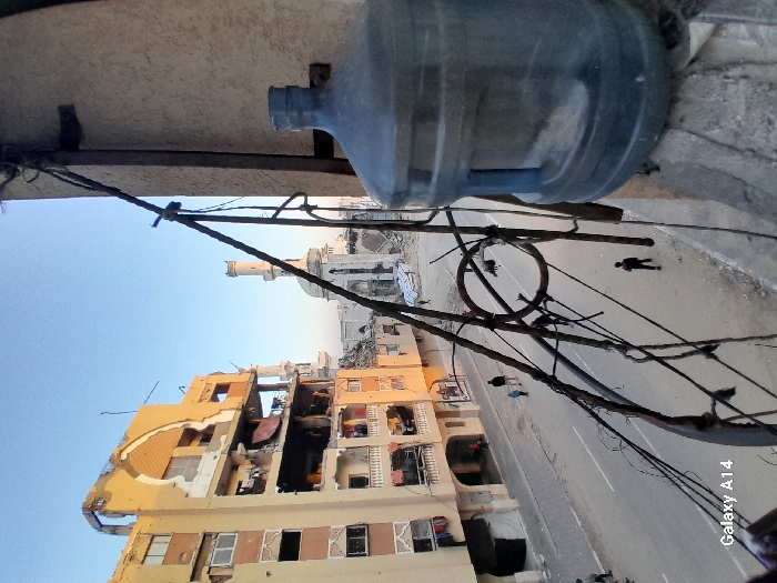 This image presents an elevated view of a heavily damaged urban street in Khan Yunis, Palestinian Territory, under a clear blue sky. In the immediate foreground on the right, a large, translucent blue plastic water dispenser bottle rests on a concrete ledge, partially obscuring the scene. Tangled dark wires and metal rods, possibly remnants of a railing or utility lines, crisscross the lower and central foreground.
To the left, a multi-story residential building exhibits extensive structural damage; large portions of its facade are missing, exposing multiple floors and their interiors, with some laundry visible hanging from balconies. Below this building, a paved street runs horizontally across the midground. Several pedestrians are present: a group of three individuals, including two adults and one child, walk away from the viewer on the paved road. Further down the street, another solitary individual walks towards the viewer, and additional figures are visible in the distance on a debris-strewn path.
In the background, a mosque with a prominent, relatively intact minaret stands amidst widespread urban destruction and piles of rubble. The surrounding environment indicates extensive damage to buildings and infrastructure.