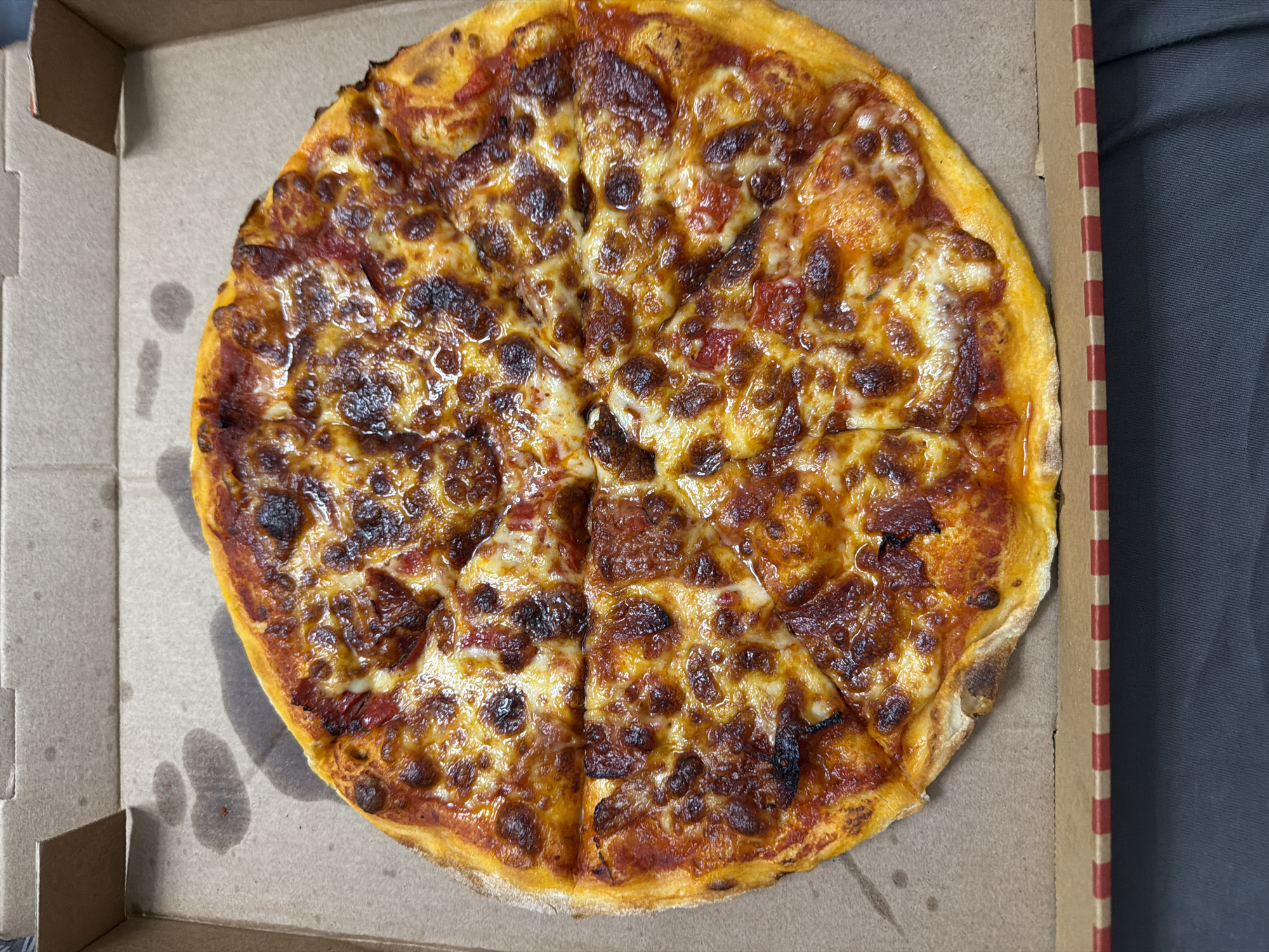 The image shows a close-up, top-down view of a whole pepperoni pizza inside a cardboard pizza box. The pizza is cut into slices and has a crispy crust, melted cheese with some browned spots, and visible pieces of pepperoni. The pizza box is open and rests on a dark gray surface. There are some water spots visible on the inside of the cardboard box near the pizza. The image does not contain any information that would allow for the determination of its location in Ruse, Bulgaria.