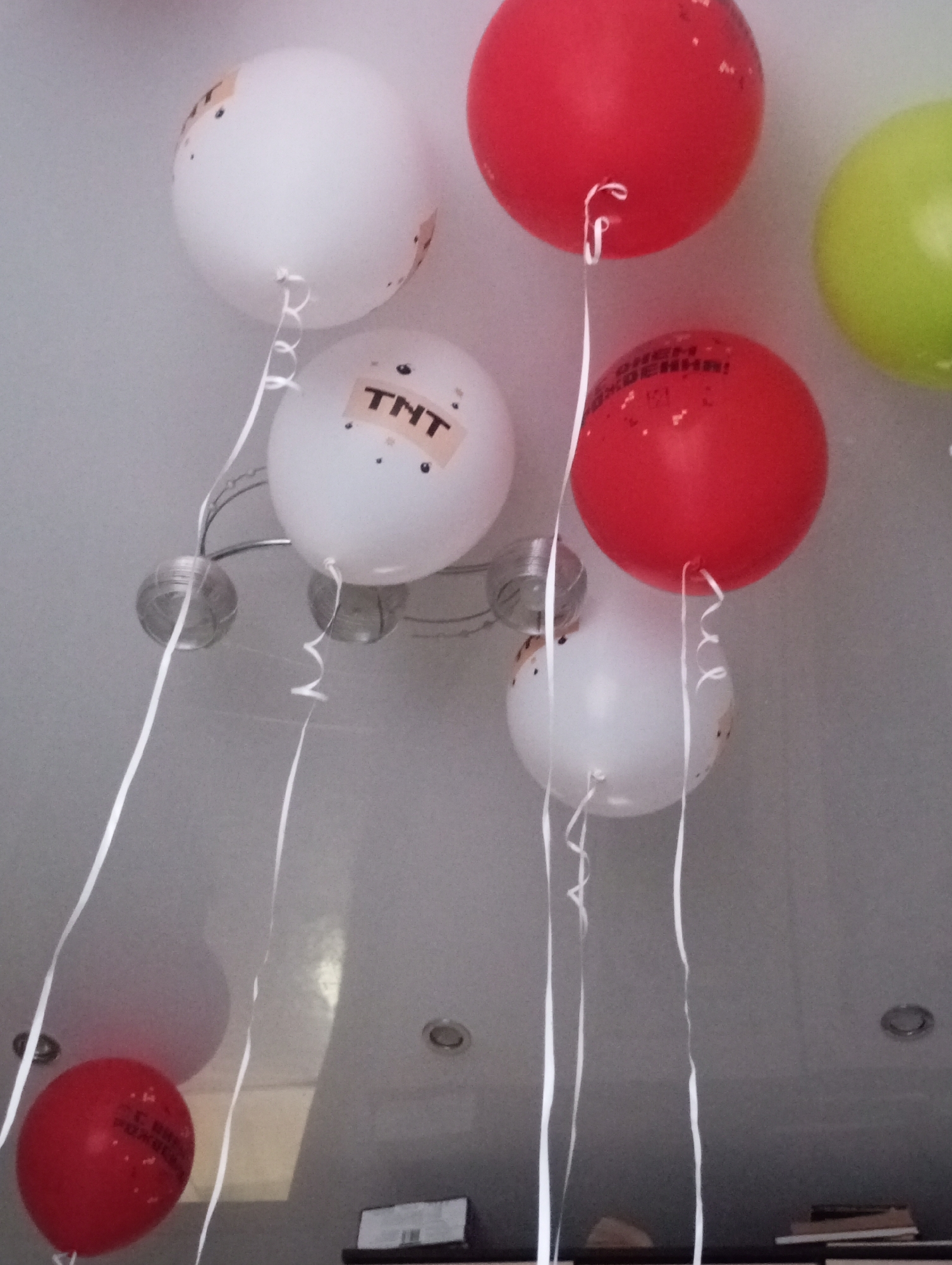 The image shows a ceiling with several balloons attached. There are white balloons with "TNT" printed on them, as well as red and green balloons. Some red balloons have text in Russian printed on them. The balloons are tied with white ribbons. In the background, a chandelier and recessed ceiling lights are visible. A shelf with books and a small box are at the bottom of the image. The city mentioned, Kamensk-Uralsky, Russia, doesn't appear to be relevant to the visual content of the image.