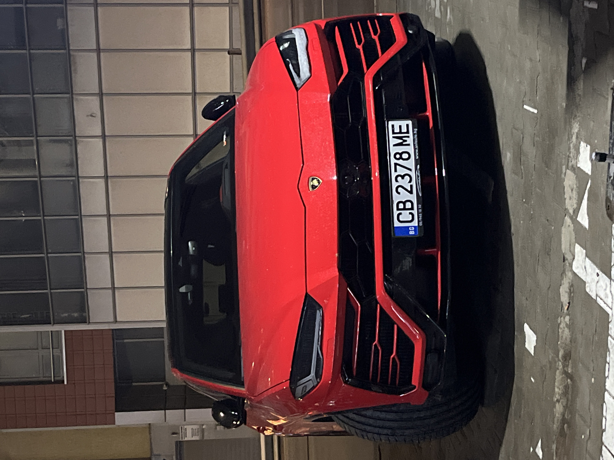 A red Lamborghini Urus is parked outdoors on a paved surface. The car's front is facing the camera, showing the license plate "CB 2378 ME," registered in Bulgaria. Based on the license plate, the location is likely in or around the city of Sofia. The background consists of a building with grid-like windows and a section of a building with brown brickwork. There are no people visible in the image. The scene appears to be at night or in low-light conditions.