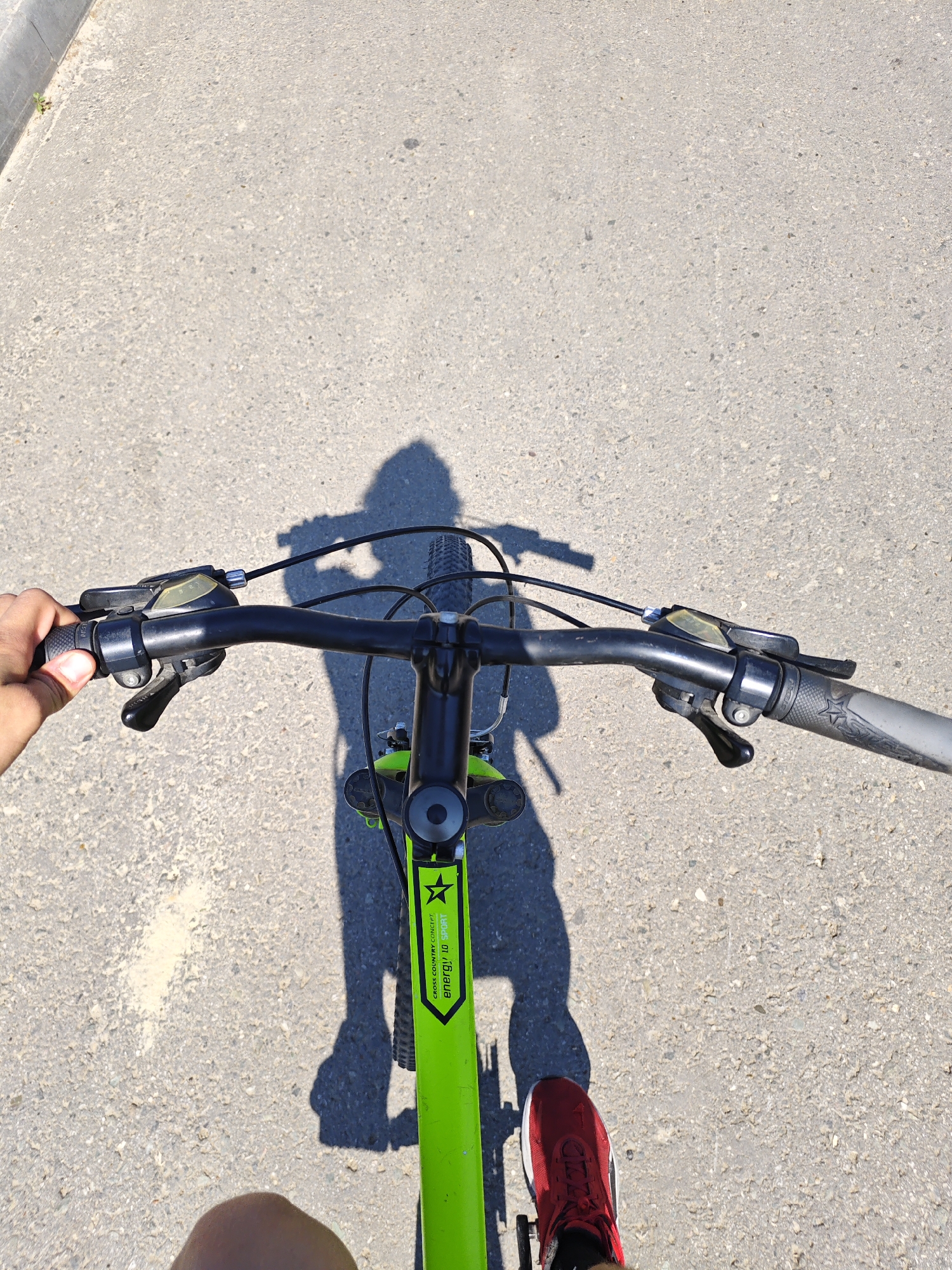 The image shows a first-person perspective of someone riding a bicycle on an asphalt surface. The handlebars of the green bicycle are prominently displayed in the foreground. A hand is visible gripping the left handlebar. The rider's right foot, wearing a red athletic shoe, is also visible. A shadow of the rider and the bicycle is cast on the ground. The environment appears to be an outdoor, paved area, possibly a street or a path. The lighting suggests it is daytime. There are no other people or significant landmarks visible in the frame.