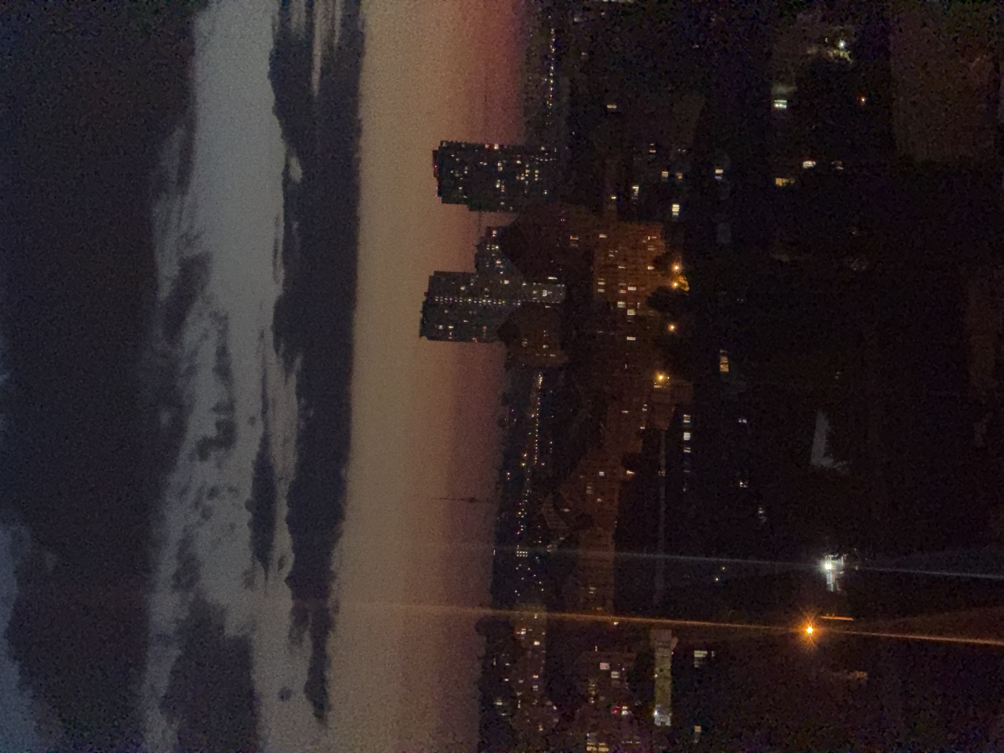 The image shows a nighttime cityscape in Kyiv, Ukraine. In the foreground, there are dark silhouettes of trees and buildings, with a lit street lamp on the left. The middle ground features a dense urban landscape with numerous lit windows in residential buildings and some darker structures. In the background, there are several tall, illuminated apartment buildings, characteristic of Kyiv's residential areas. Further in the distance, a slender tower, possibly a communication tower, is visible against the dusky sky. The sky itself is a mix of dark, heavy clouds and a gradient of fading light from a sunset or twilight, casting an orange and purple hue. The overall impression is of a quiet, developing city at dusk or after dark.