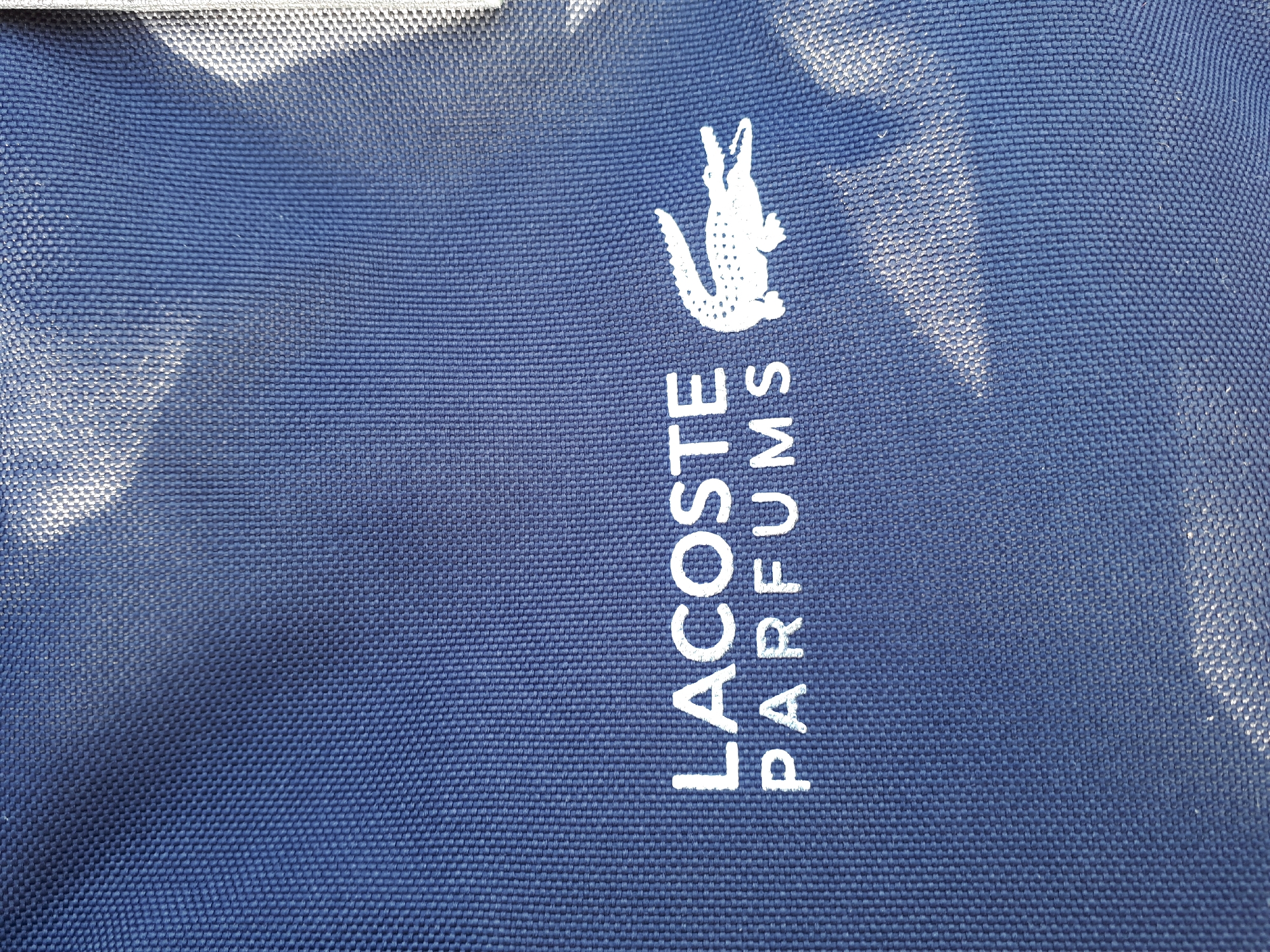 A close-up, angled shot shows a dark blue fabric with a white logo. The logo consists of the words "LACOSTE" and "PARFUMS" printed in white capital letters, positioned one above the other. To the right of the text is a white graphic of a crocodile. The fabric has a visible woven texture, and a bright light casts a shadow on the right side of the image, illuminating the woven threads in that area. The image is taken from an unknown location in an unknown city.
