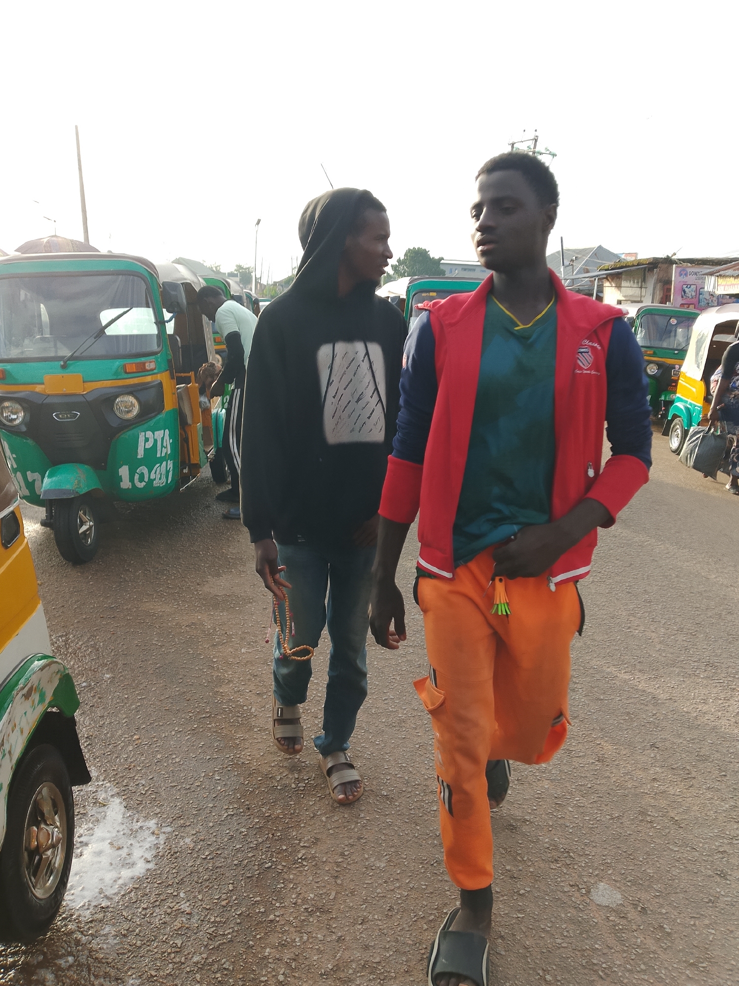 Two young men are walking along a dirt road in Jos, Nigeria. The man in the foreground is wearing a red jacket over a green shirt and bright orange pants, and he is walking towards the right of the frame. He has dark skin and short black hair, and he is looking slightly to his left. The man behind him is wearing a black hooded sweatshirt and blue jeans, and he is walking towards the left of the frame, looking at the man in front of him. He has dark skin and short black hair, and he is holding a string of beads in his right hand.
Several auto-rickshaws, locally known as "Keke Napeps," are parked along the side of the road. One of the Keke Napeps is bright green with yellow accents and has "PTA 1041" written on its side. Another Keke Napep in the background is yellow and green. The road surface is a mix of gravel and dirt, and there are some small puddles of water on the ground. The sky is bright and appears to be late afternoon or early morning. In the background, there are some buildings with corrugated metal roofs, and some trees. The overall scene suggests a bustling local transportation hub or a market area.