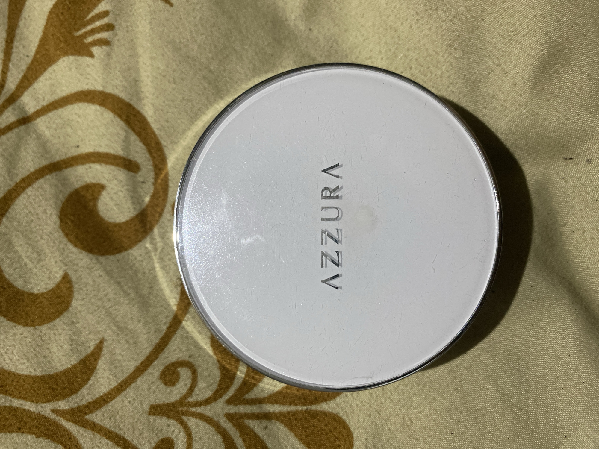 A white, round cosmetic compact with silver trim is centered in the frame, resting on a textured fabric with a tan and brown floral pattern. The brand name "AZZURA" is embossed in silver lettering across the top surface of the compact. The fabric appears to be a tablecloth or bedspread, possibly located in Ta', Indonesia. The lighting suggests an indoor setting.