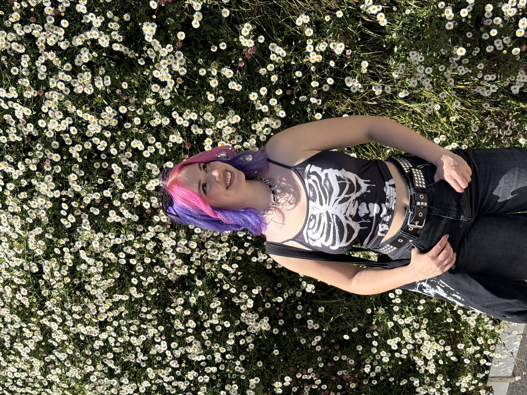 A young woman is lying in a field of small, white daisies, likely in an outdoor area. She has dyed hair in shades of pink and purple and wears a black tank top with a white skeleton rib cage design. Her jeans are black, and she wears two studded belts. The environment is sunny, suggesting it could be a park or garden where wildflowers are growing. The location is not discernible as being in Madrid, Spain without additional context or visual cues specific to that city.