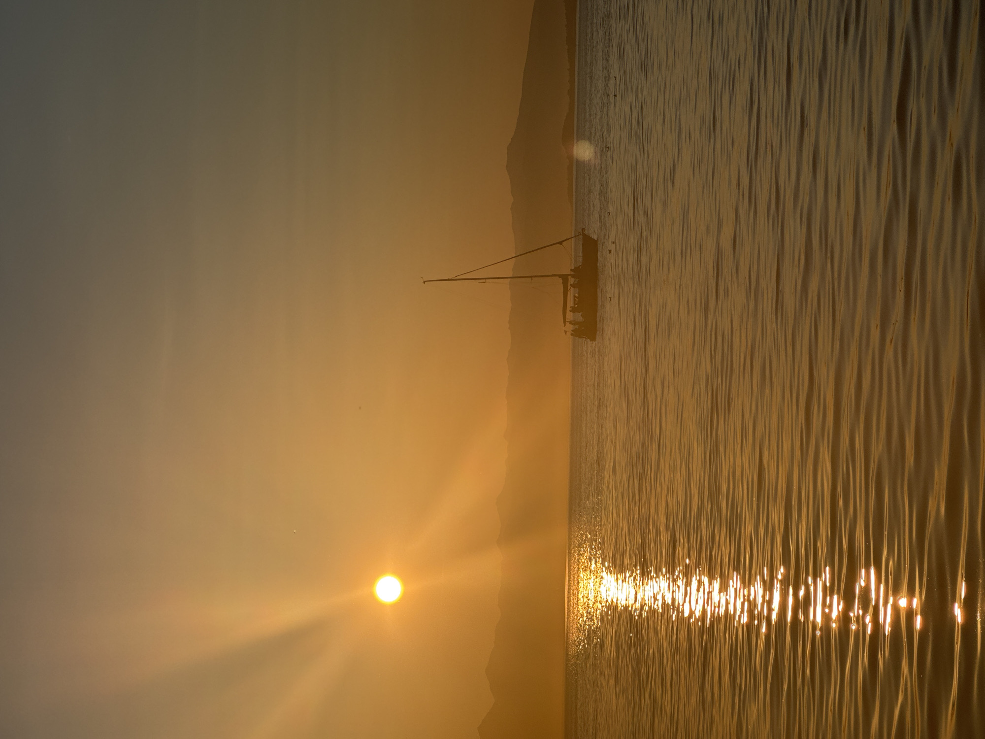 The image depicts a tranquil seascape at sunset, with the sun casting a warm golden glow across the water. In the distance, silhouetted mountains form the horizon line. A sailboat with several people on board is visible in the middle ground, gently gliding across the calm sea. The sun's rays are piercing through the atmosphere, creating a dramatic sunburst effect. The water is smooth with gentle ripples, reflecting the vibrant colors of the sunset. The scene is serene and picturesque, evoking a sense of peace and natural beauty. The image does not provide any specific clues to pinpoint the location within Petropavlovsk-Kamchatsky, Russia.