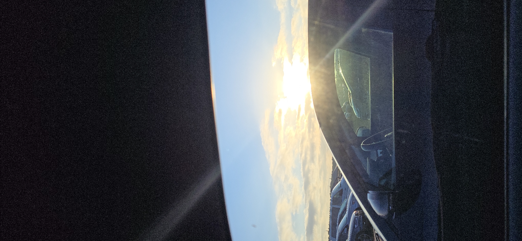 The image is a photograph taken from inside a vehicle looking out at a parking lot with a bright sun setting or rising in the sky. The foreground shows the interior of a car, with the dashboard and a portion of the driver's side window visible. Outside the window, several cars are parked in rows under a sky filled with clouds. The sun is a bright, intense light source, casting rays across the scene. The overall lighting suggests it is either early morning or late evening. The location in Neustadt-Glewe, Germany, is not visually discernible from the image itself.