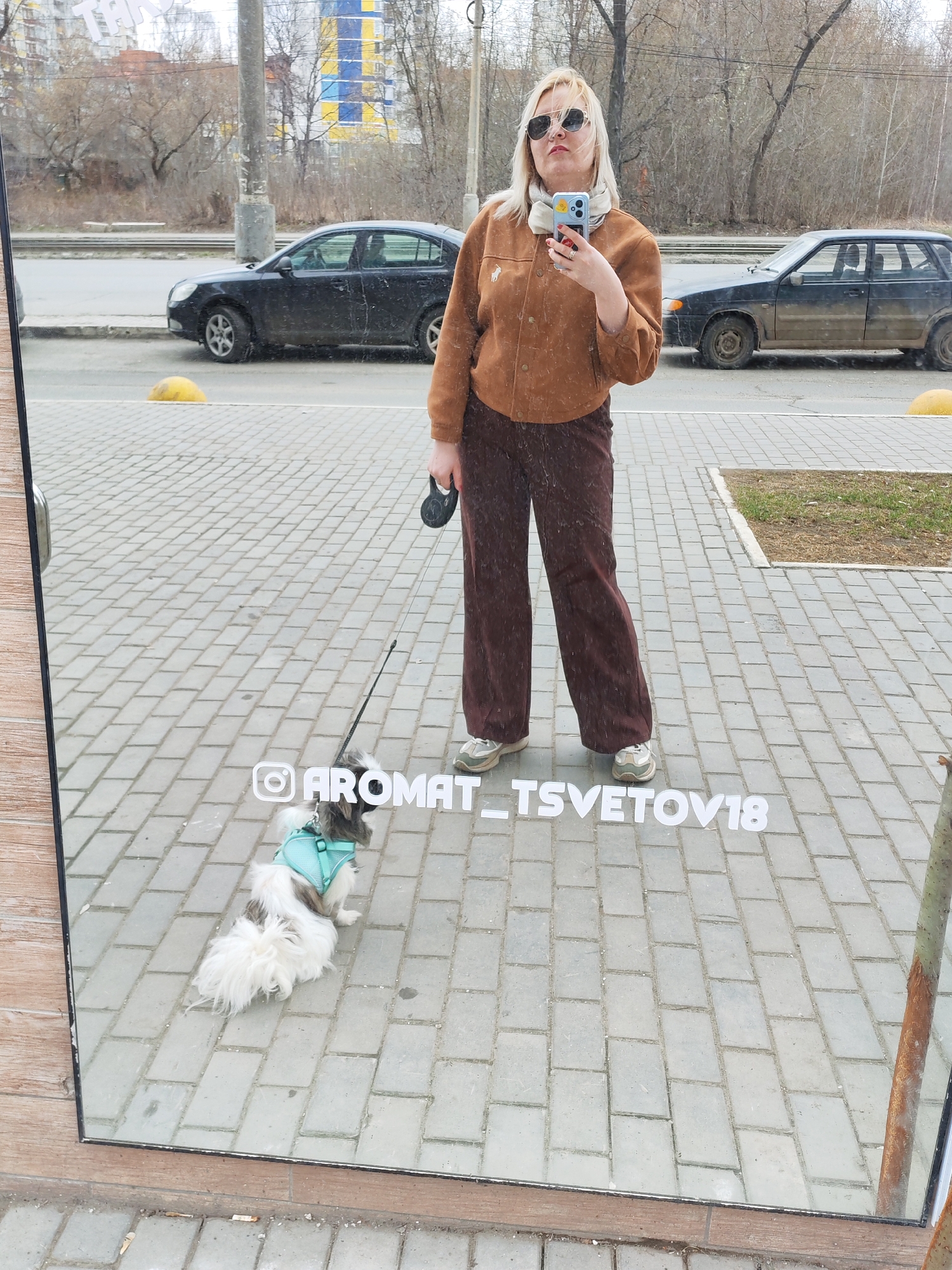 A woman in sunglasses stands on a paved sidewalk, reflecting in a large mirror with a dog on a leash. She wears a brown jacket and trousers, holding a phone with a beige scarf around her neck. The dog is small, with a light blue harness. The username "@AROMAT_TSVETOV18" is overlaid on the sidewalk in the mirror. Cars are visible on the street behind, and buildings can be seen in the background. The scene is Izhevsk, Russia.