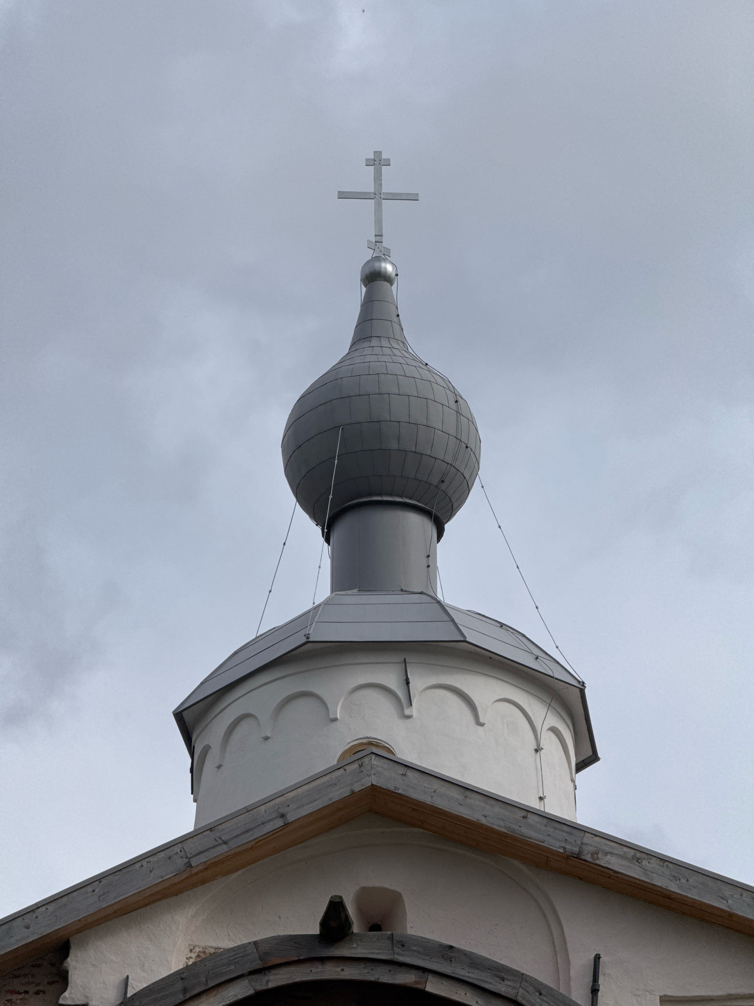 The image shows the dome of a white building topped with a silver cross against a cloudy grey sky. The dome is dark grey and bulbous, with a lighter grey band encircling its base. Thin wires are attached to the dome and extend downwards. Below the dome is a white structure with arched architectural details. A wooden roof overhangs this structure. The architectural style suggests it is likely a religious building, possibly an Orthodox church. Given the architectural style and location, this could be a part of the Saint Sophia Cathedral or another historical church in Veliky Novgorod, Russia. There are no people visible in the image.